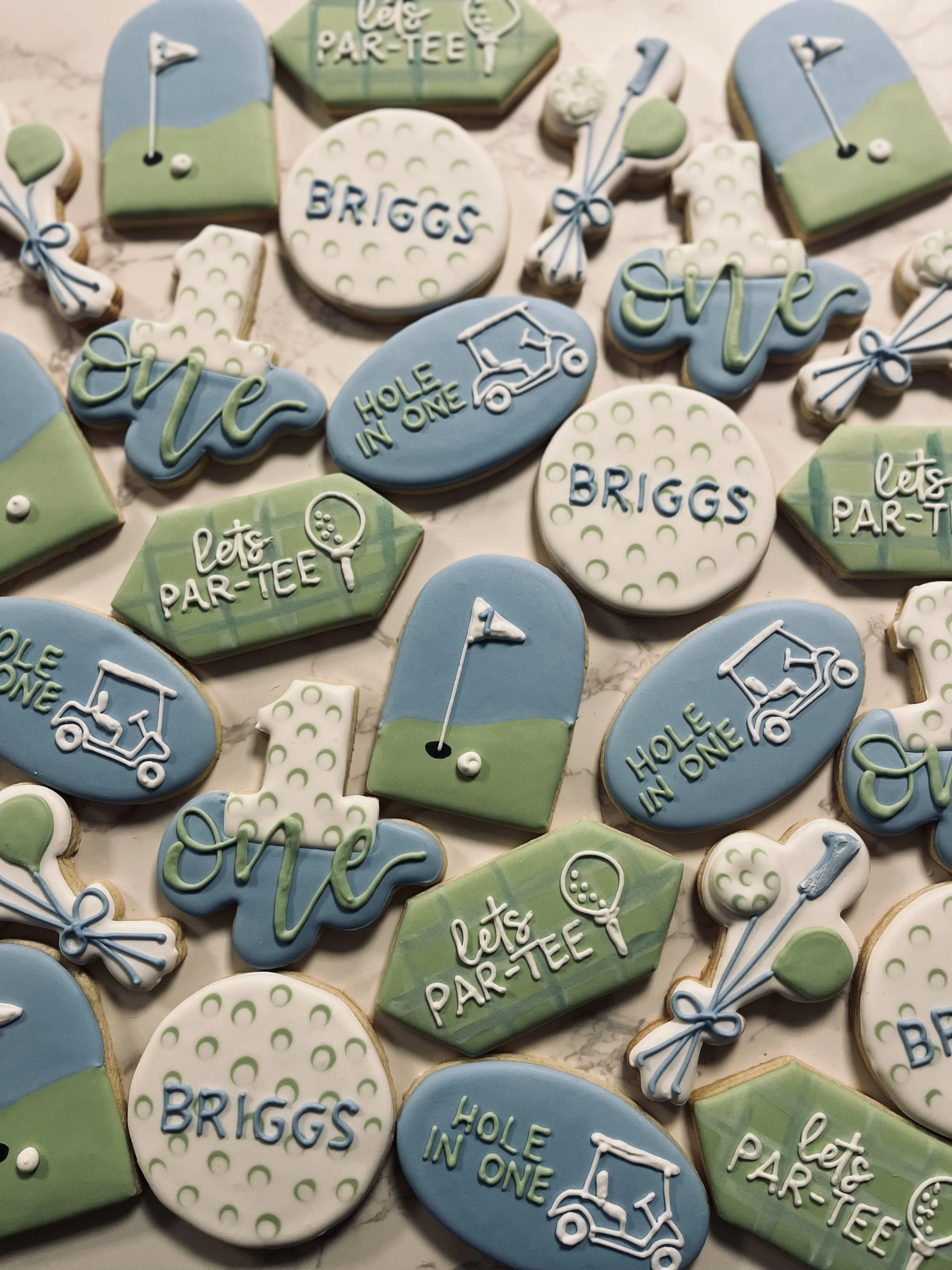 Golf-themed decorated cookies with phrases and symbols like golf flags, golf carts, and phrases such as 'let's par-tee,' 'hole in one,' and 'bring #Briggs' in green, blue, and white icing.