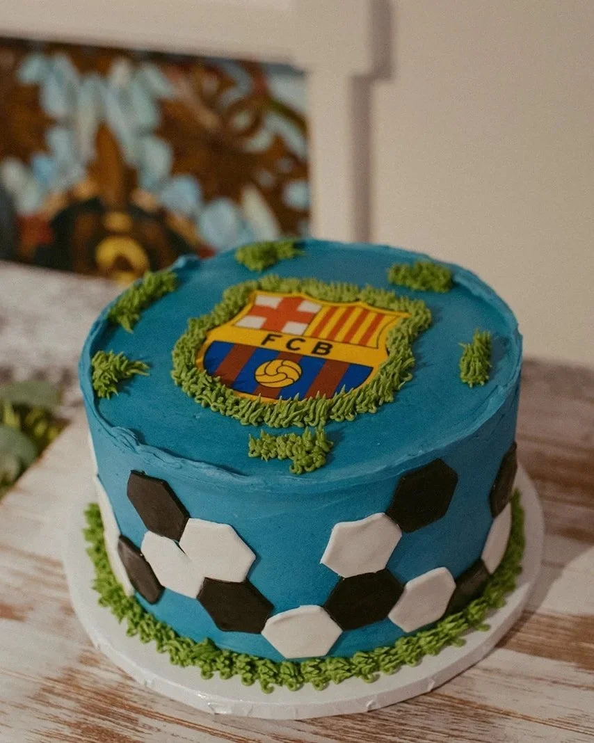 A birthday cake decorated with FC Barcelona soccer team emblem on top and black and white soccer ball pattern around the sides.