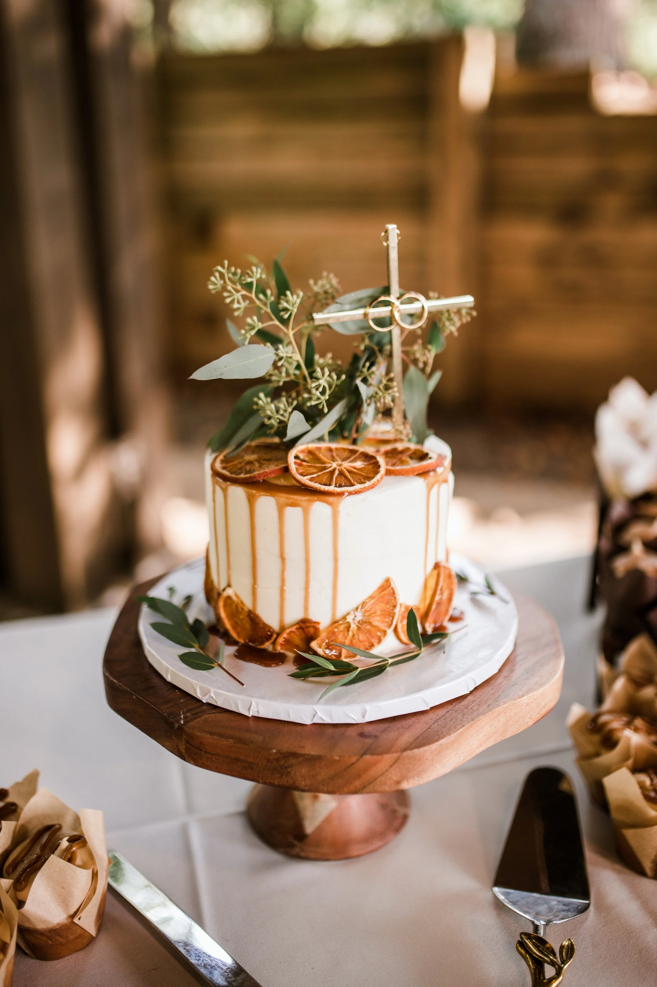 A decorative cake with a caramel drip, dried orange slices, eucalyptus leaves, and a metallic ring cross topper on a wooden cake stand at a celebration.