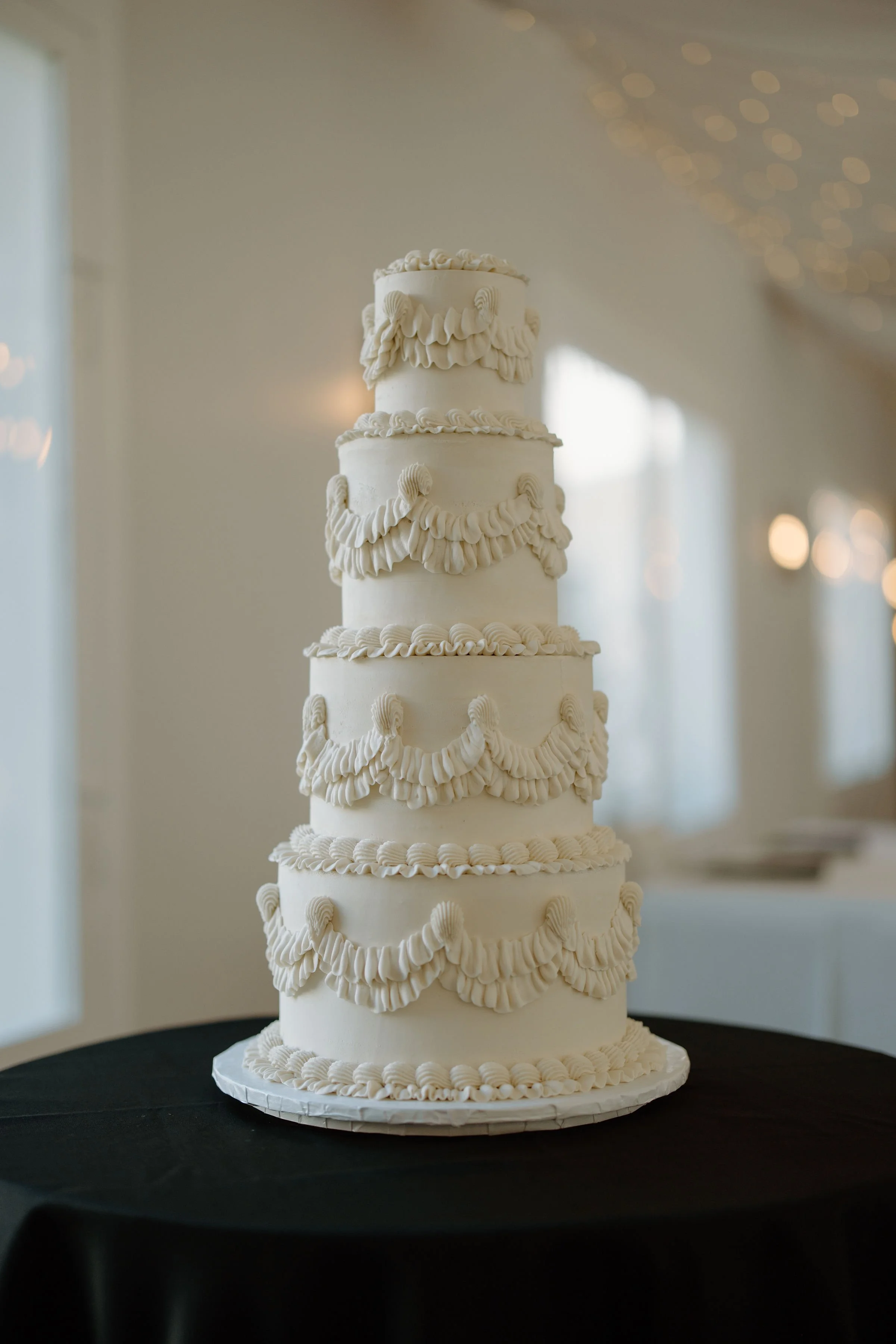 A tall, white, multi-tiered wedding cake with decorative piping and swags, placed on a black tablecloth.