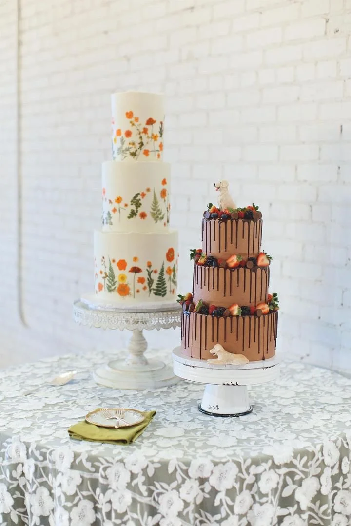 Two decorated cakes on stands, one with floral designs and another chocolate with fruit and dog figurines, on a lace-covered table.