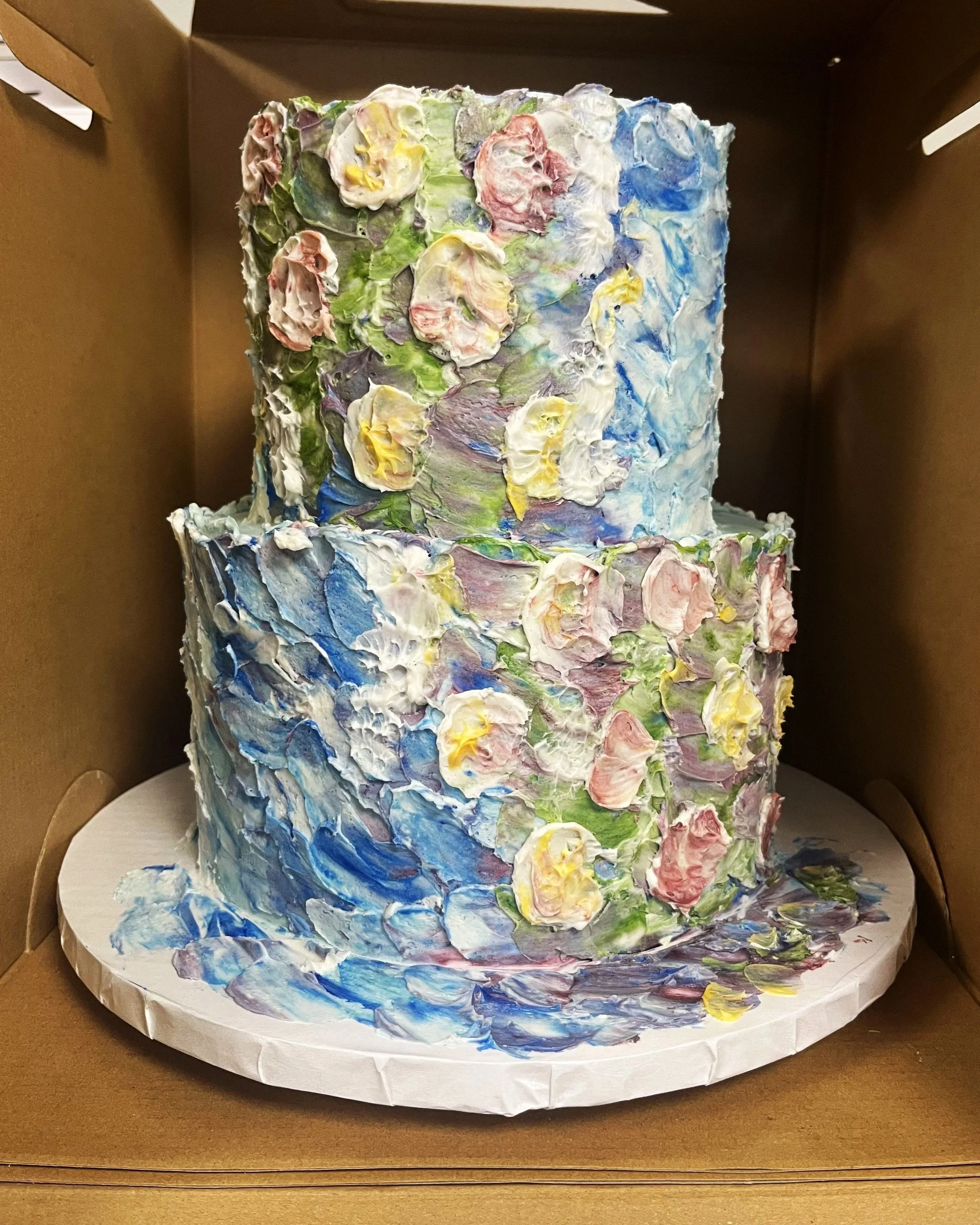 Two-tiered cake decorated with a thick, impressionist-style buttercream floral design in pastel colors, placed inside a brown cardboard box.