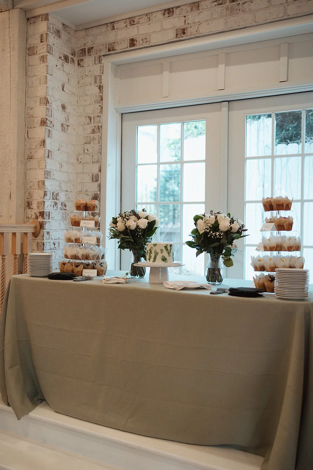 A dessert table decorated with white flowers and cupcakes, set against large window doors with a white brick wall backdrop.