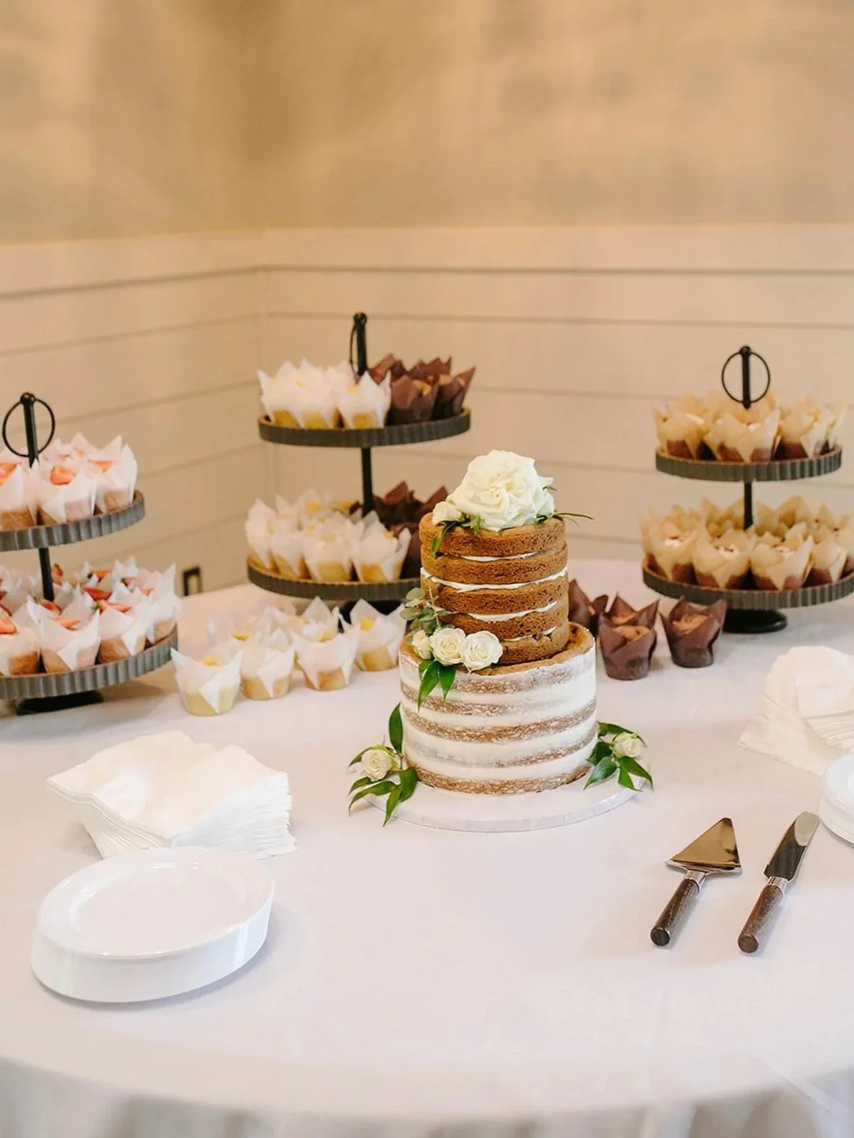 A two-tiered rustic wedding cake decorated with white roses and greenery, placed on a white table with assorted cupcakes on tiered stands in the background.