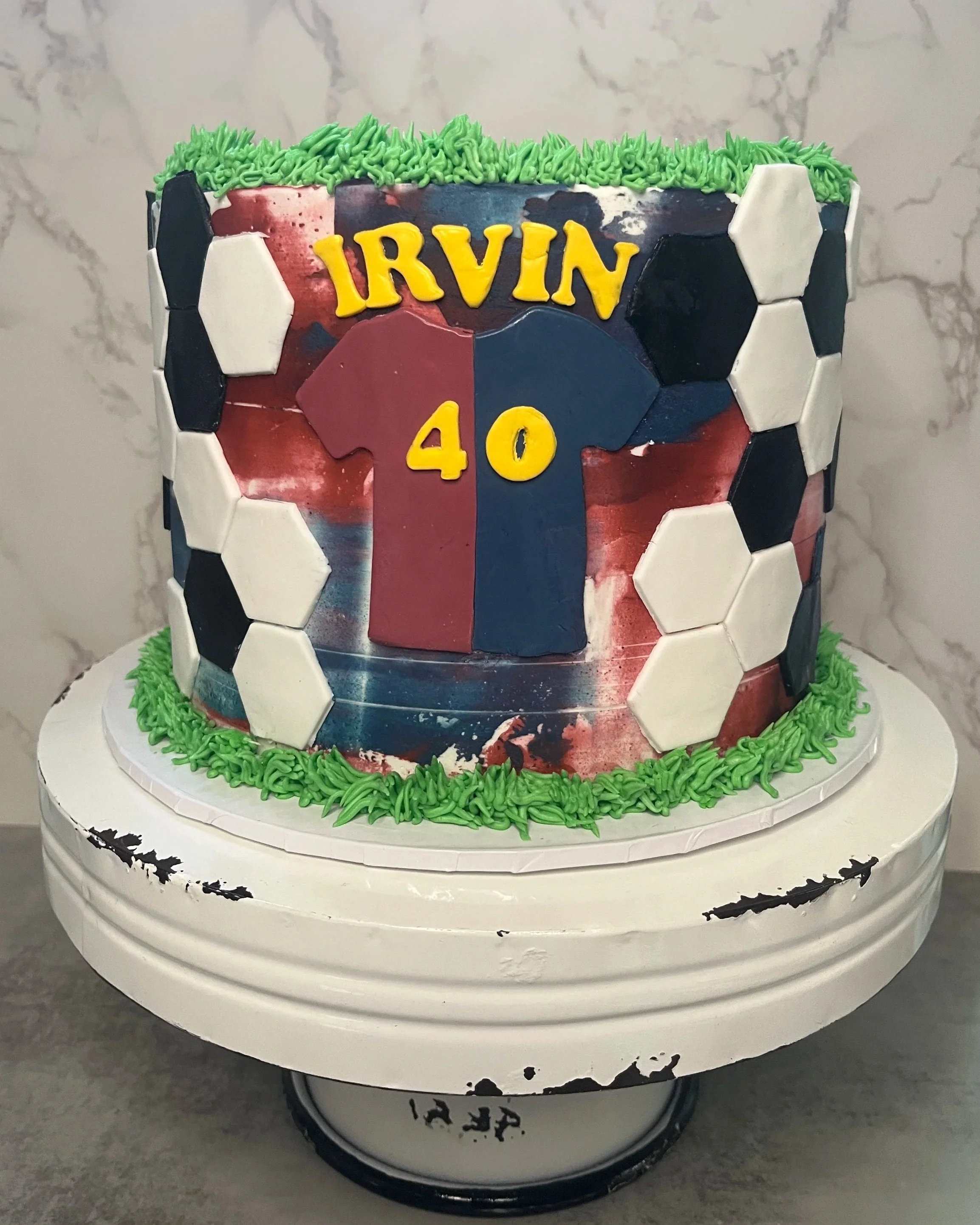 A soccer-themed birthday cake with the name Irvin, age 40, decorated with soccer ball patterns and a jersey design, topped with green piped frosting, on a white cake stand.