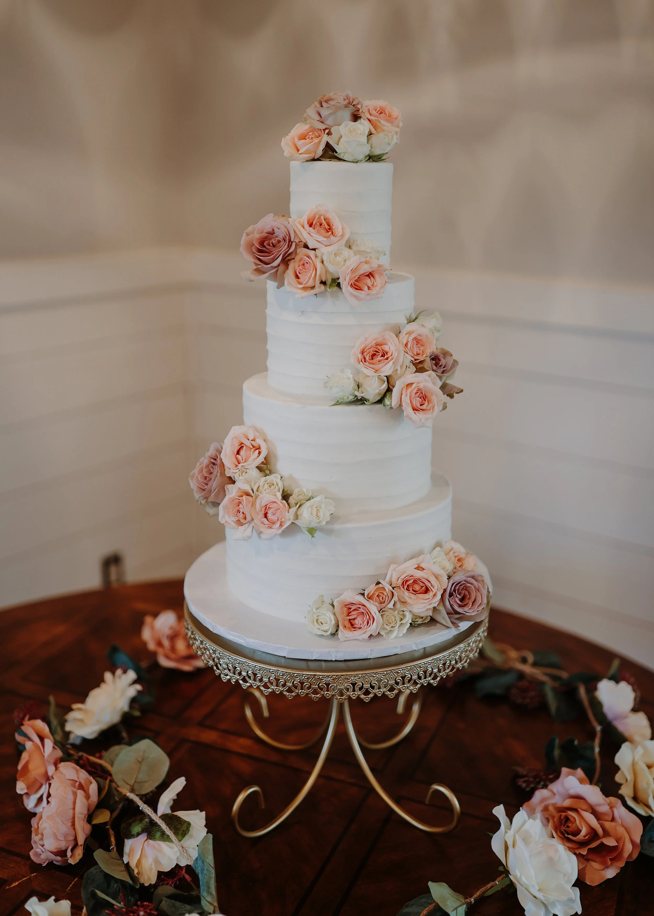 Four-tier white wedding cake decorated with pink and white roses, placed on a decorative silver stand on a wooden table surrounded by scattered roses.