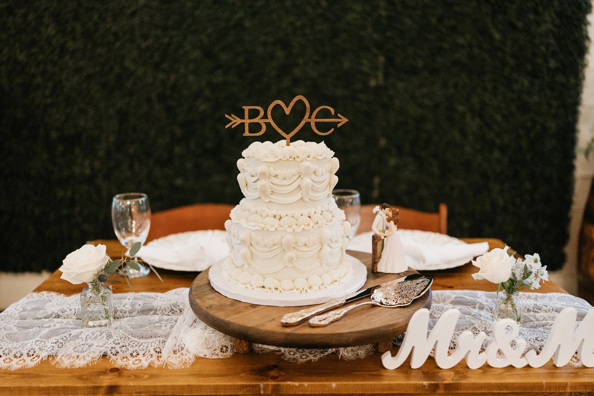 A multi-tiered white wedding cake with decorative icing is displayed on a wooden cake stand, decorated with a cake topper that spells 'LOVE' with a heart and arrows, surrounded by floral arrangements on a lace-covered table.