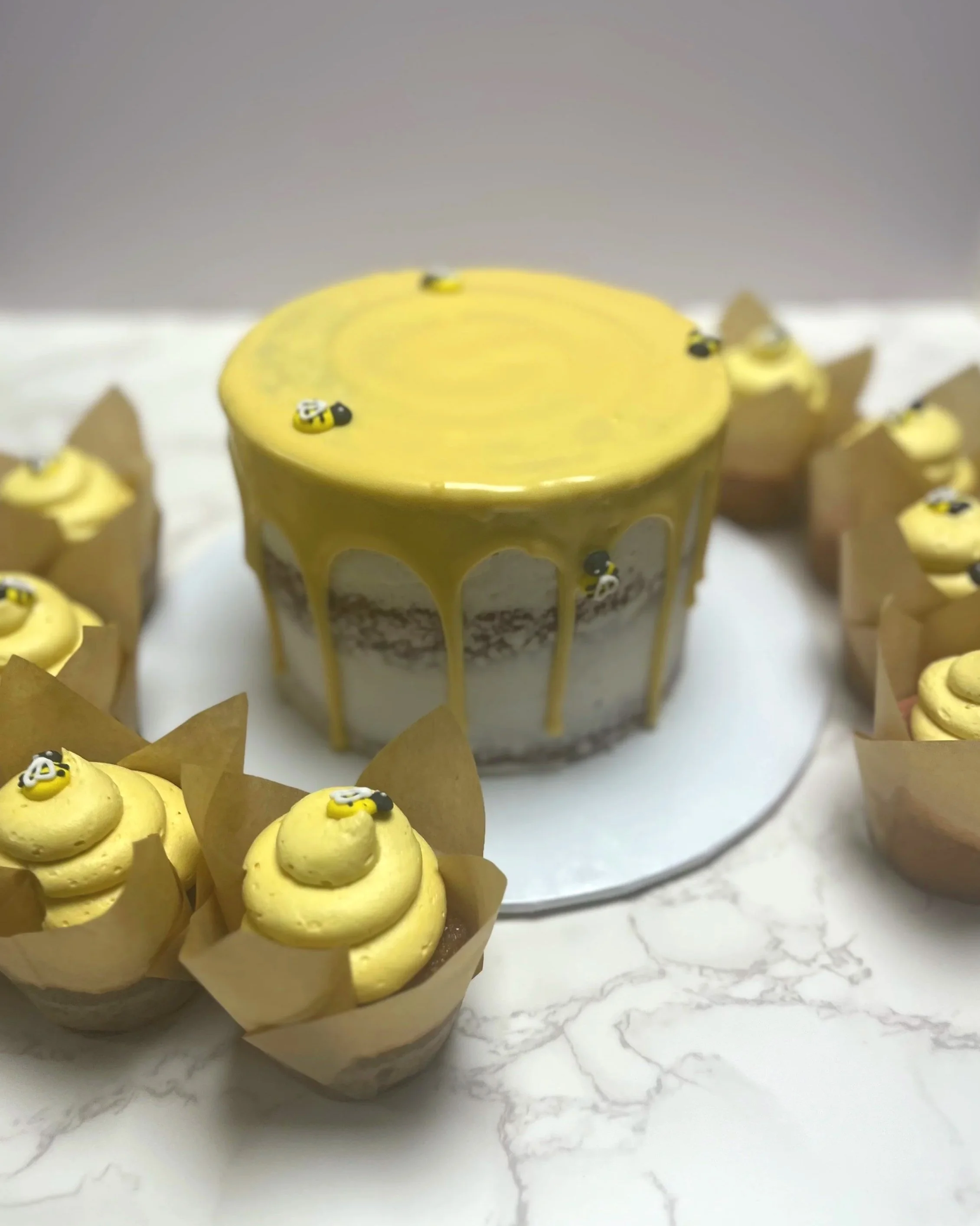 A cake with yellow glaze and small bees decoration surrounded by cupcakes with yellow frosting and bee decorations on a white surface.
