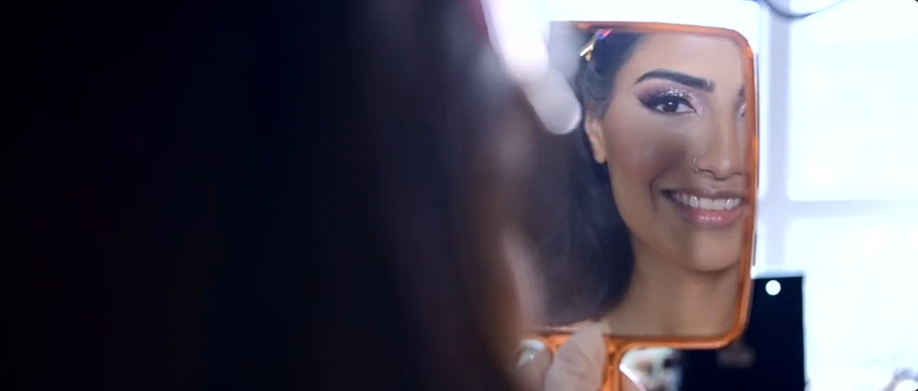A woman with dark hair and makeup smiling at herself in a mirror while applying makeup.