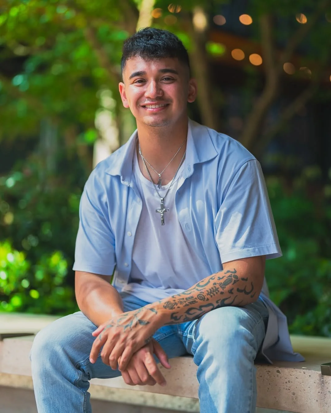 A young man with short dark hair, smiling, wearing a light blue shirt over a white T-shirt, accessorized with necklaces and tattoos on his arm, sitting outdoors on a wooden bench with a green, leafy background.