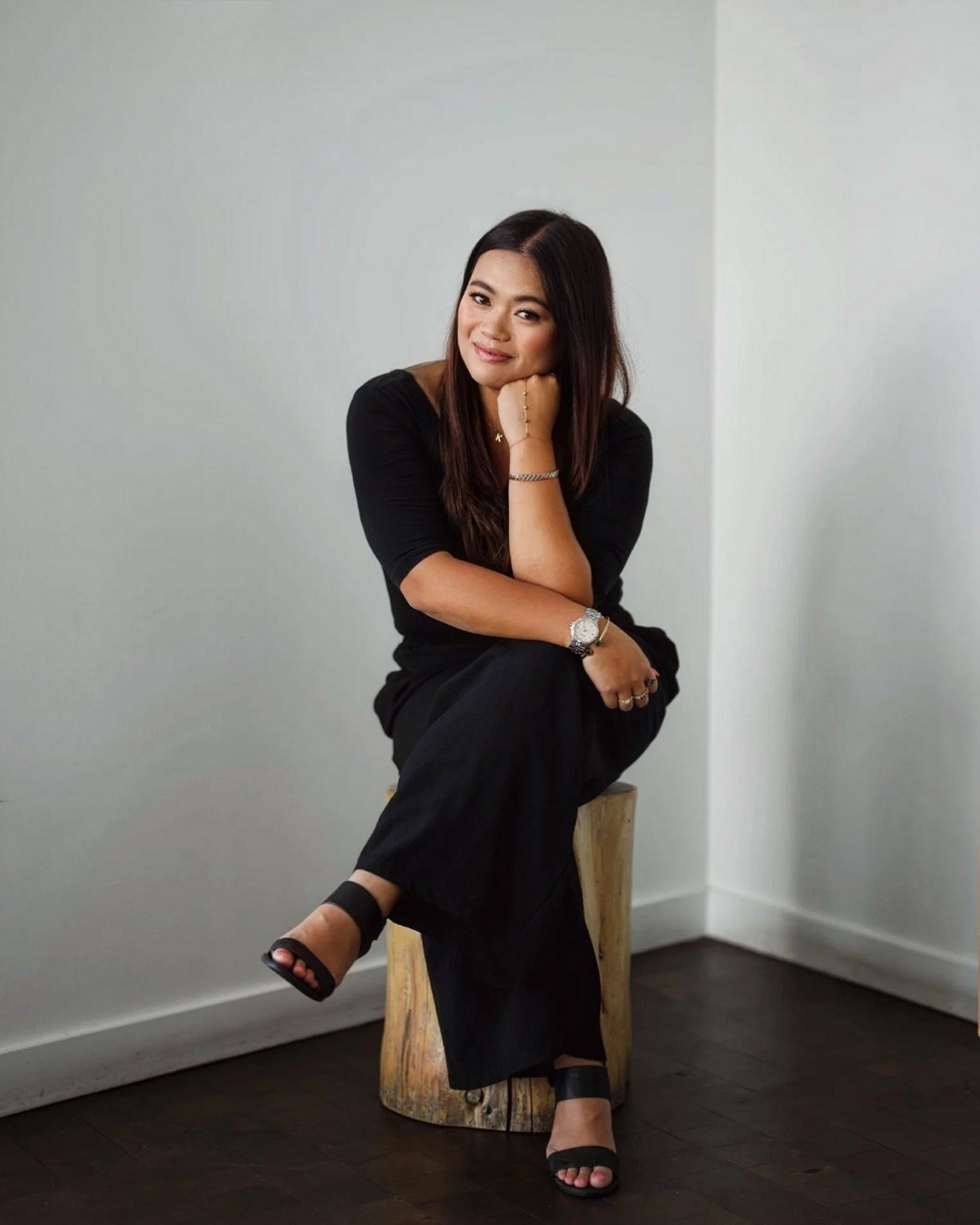 A woman with long dark hair, wearing a black dress and black high-heeled sandals, sitting on a wooden stool in a corner of a room with plain white walls, smiling gently and resting her chin on her hand.