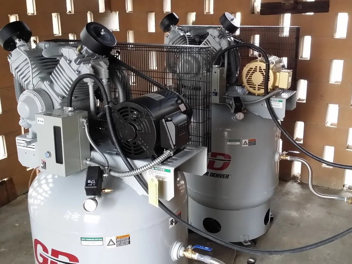 Two Gardner Denver 7.5 HP air compressor, connected by Parker Pushlok hoses, placed on a concrete floor against a concrete square cutouts enclosure, completed by The Compressor Guy, LLC