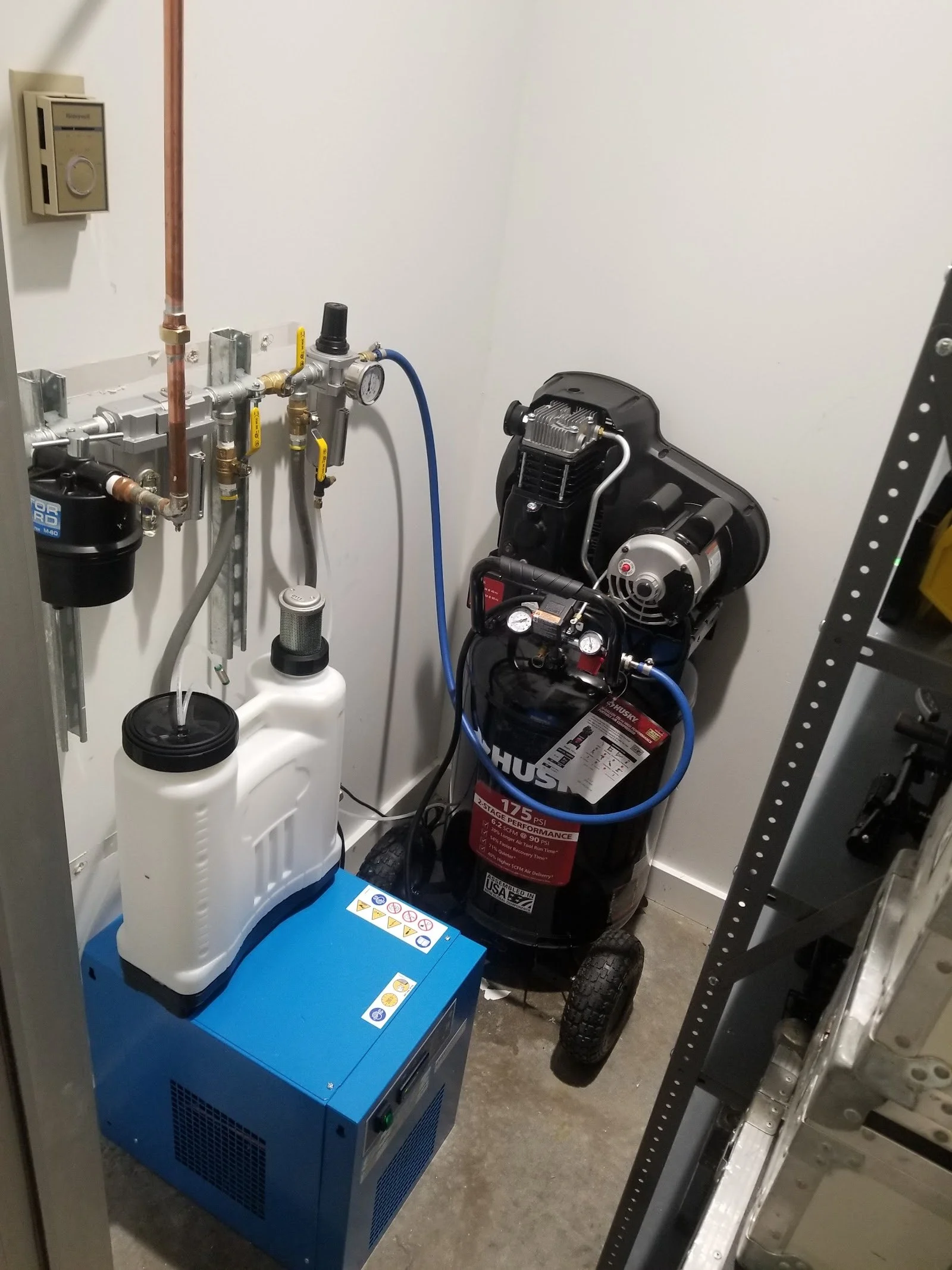 A small utility room with a blue refrigerated air dyer, white condensate collection vessel, a portable Husky air compressor, and various bypass plumbing, filtration, and electrical connections on the wall installed by The Compressor Guy, LLC.