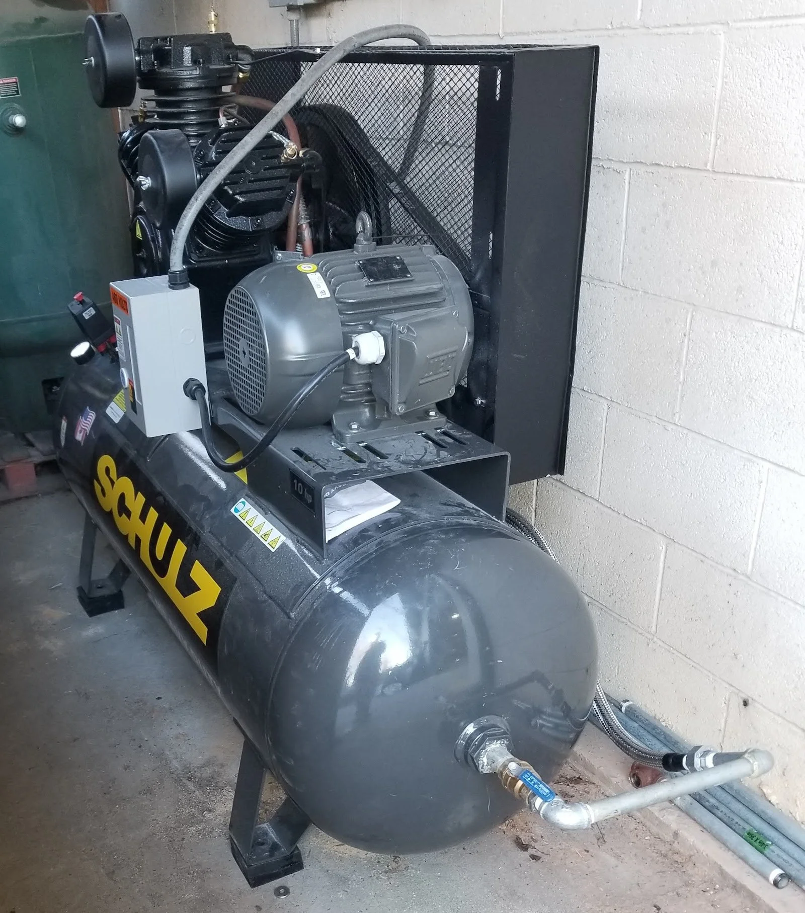 A Shultz 15 HP two stage air compressor with a mounted fan guard, connected to rigid plumbing and pipes, sitting on a concrete floor repaired by The Compressor Guy, LLC.