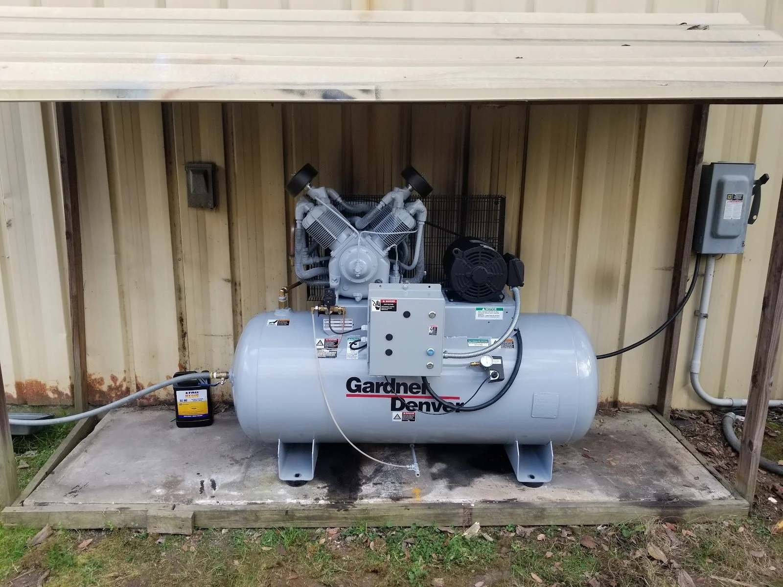 A Gardner Denver 7.5 HP air compressor installed against a metal siding wall, connected by Parker Pushlok hoses and electrical SOOW cord to the electrical panel, situated on a concrete platform installed by The Compressor Guy, LLC.