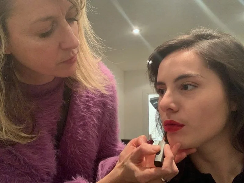 A makeup artist applies red lipstick to a woman in a salon or studio setting.