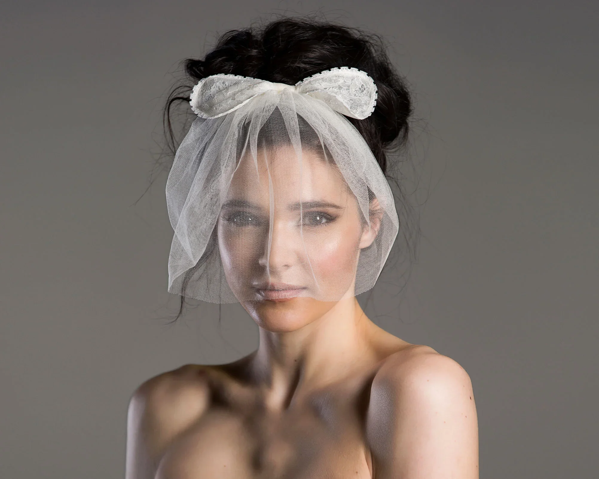 A woman with dark hair styled in an updo, wearing a white lace headband and veil, looking at the camera with a neutral expression against a plain gray background.