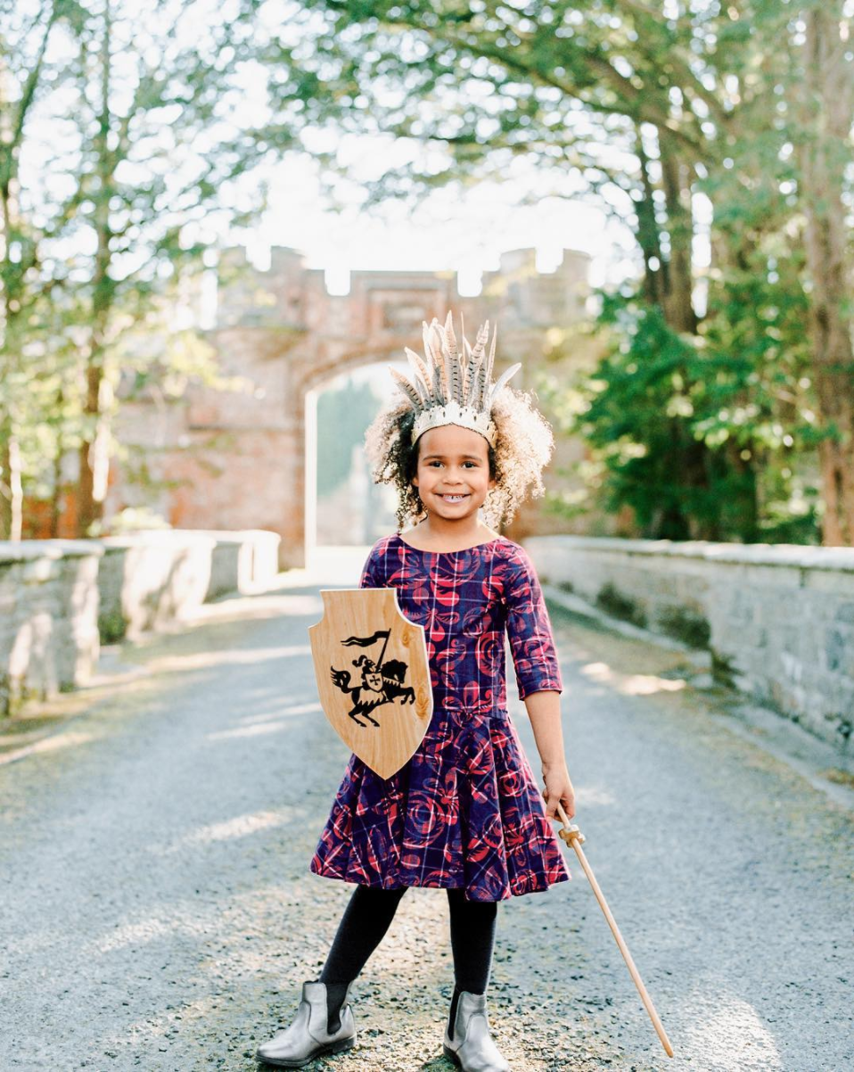 Tea Collection 20217 - young model wearing a handmade crown of feathers, holding a handmade shield, dressed in a bold patterned dress and silver Chelsea boots.