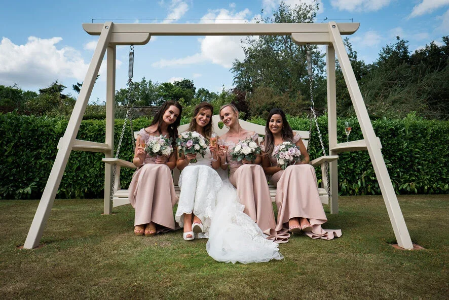 Bridal hair and makeup by Julie McGuire, Edinburgh, for your Bridesmaids. Four women sitting on a large outdoor swing, with the bride in a white wedding dress and three bridesmaids in matching pink dresses, smiling and holding bouquets of flowers.