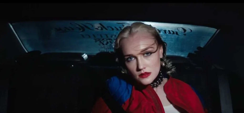 Hair and makeup for music video - The Ninth Wave Scotland - A woman with blonde hair, red lipstick, and a red jacket sitting in the back seat of a car, looking directly at the camera with a serious expression.
