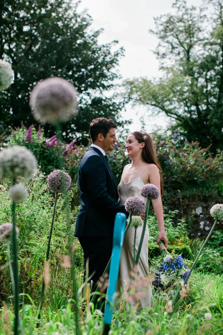 A couple dressed in wedding attire standing in a lush garden with purple and white flowers, sharing a tender moment and smiling at each other.