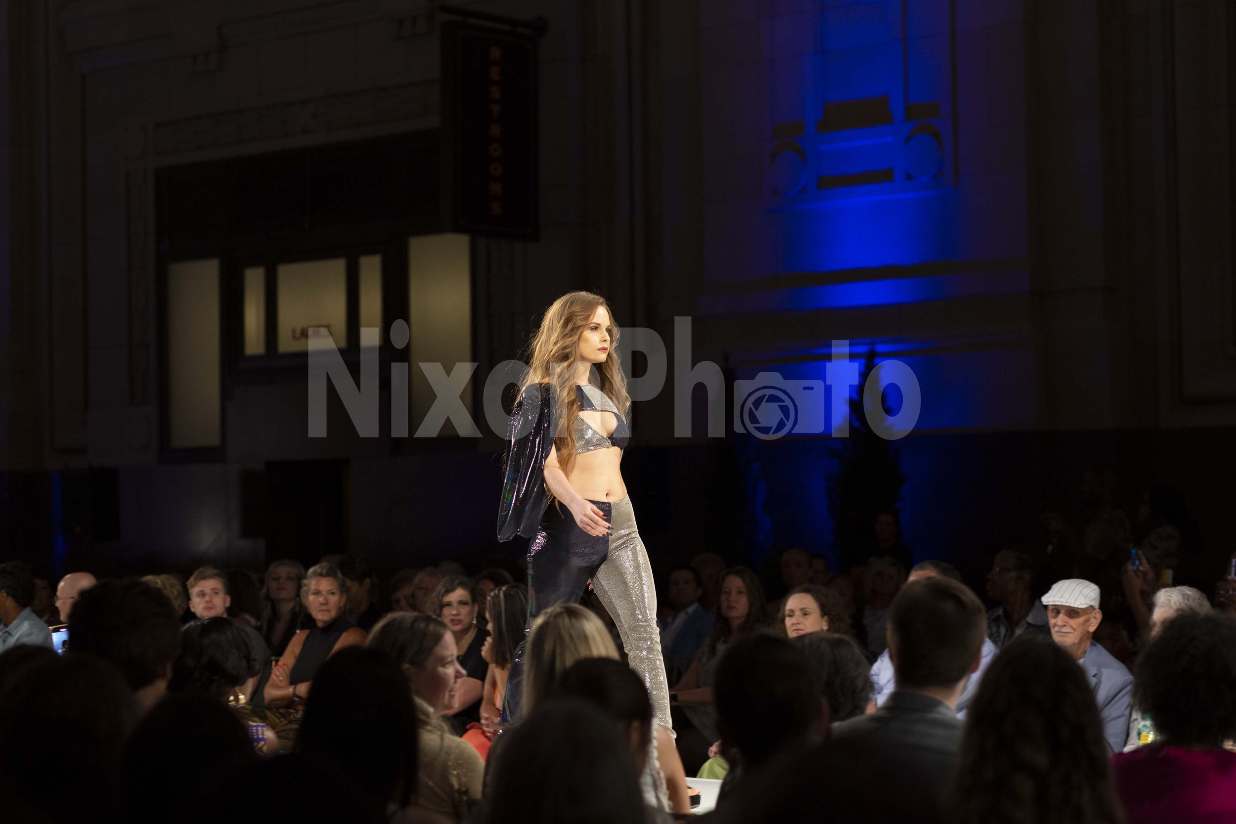 Fashion show with a model walking the runway wearing a metallic outfit, surrounded by an audience in a dimly lit venue.