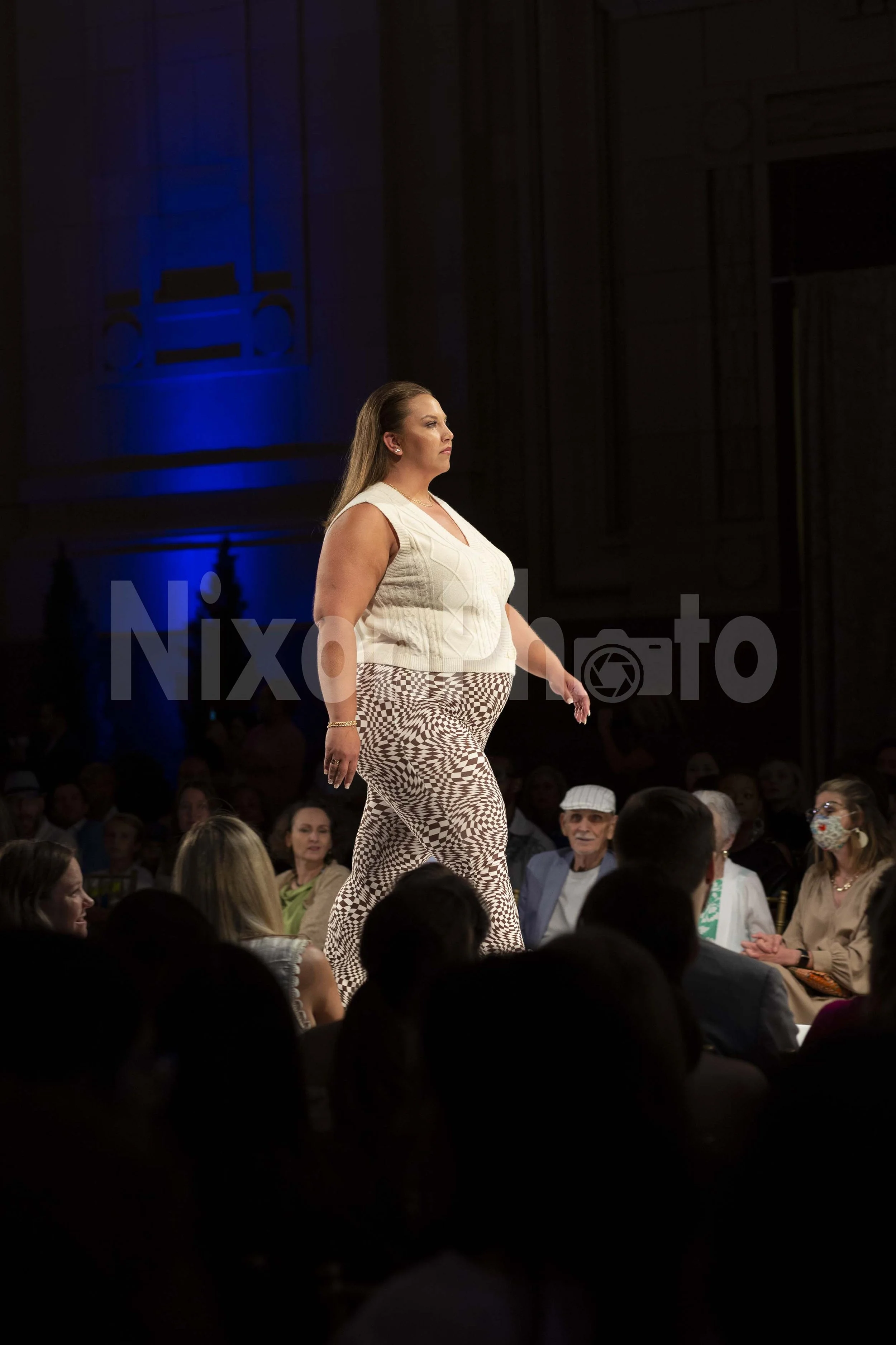 Plus-size model walking on a fashion runway wearing a white sleeveless top and patterned pants in front of an audience.