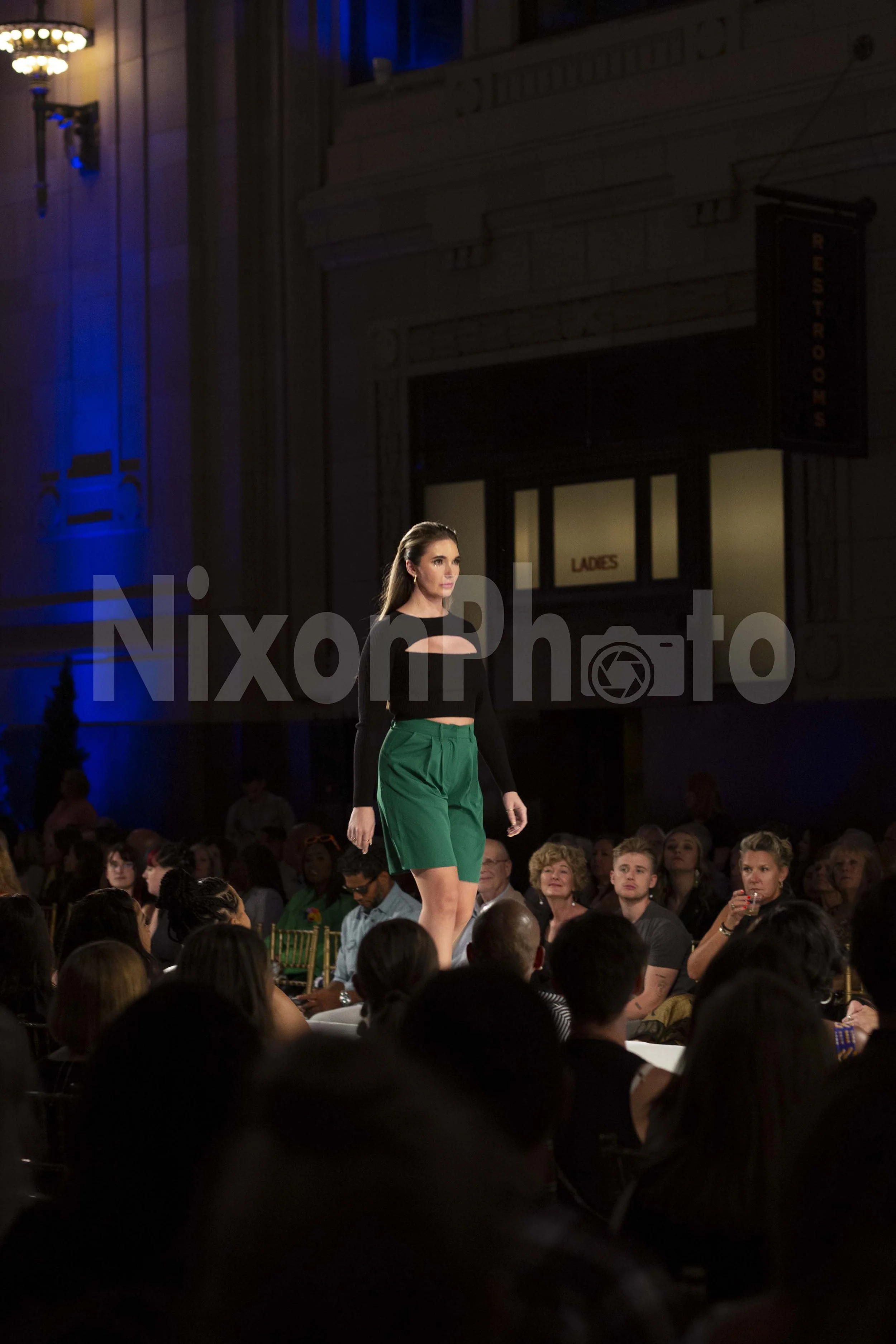 Fashion model walking on runway in black top and green skirt at nighttime event.