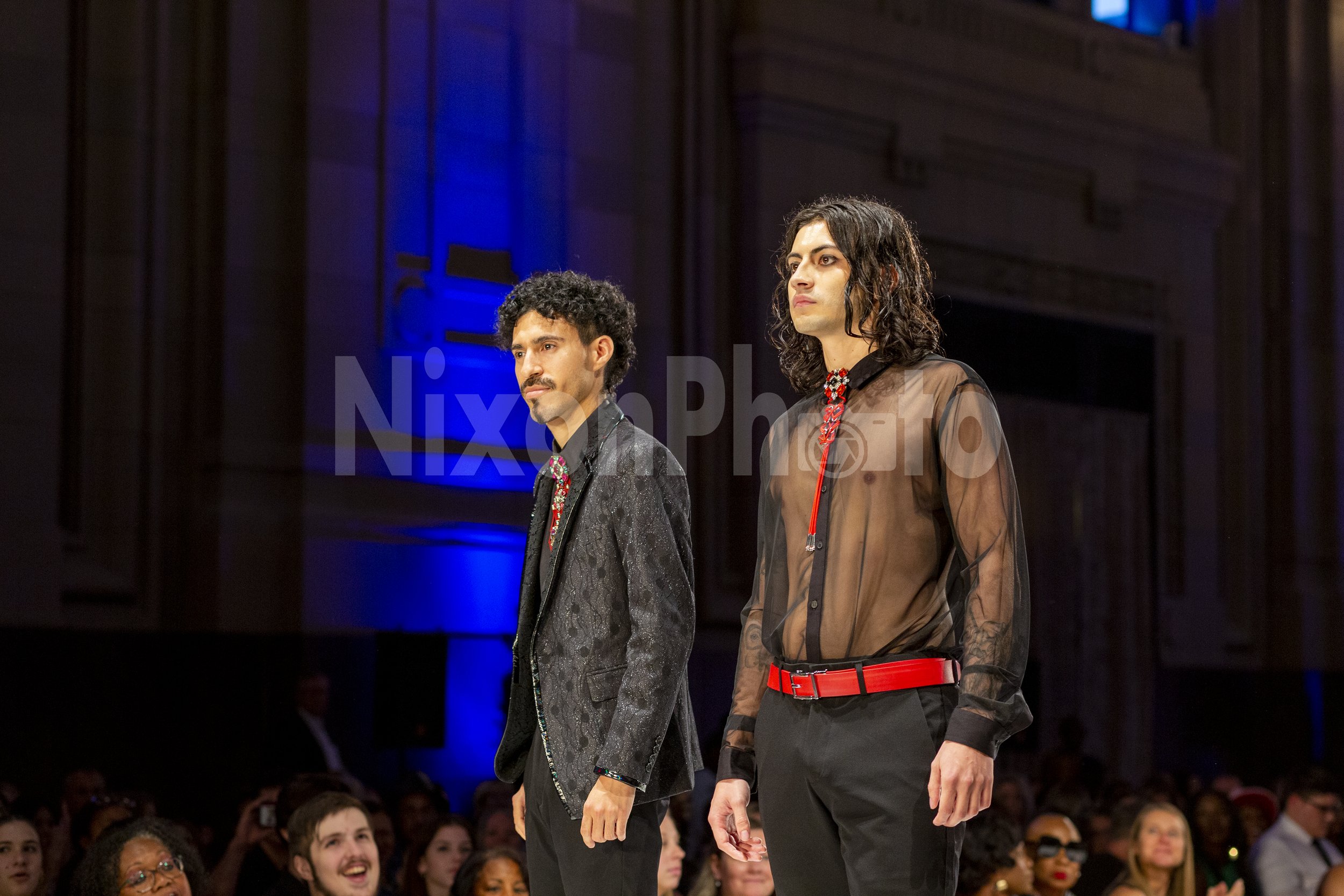 Two male models on a runway; one wears a patterned suit and the other a sheer shirt with red accents.