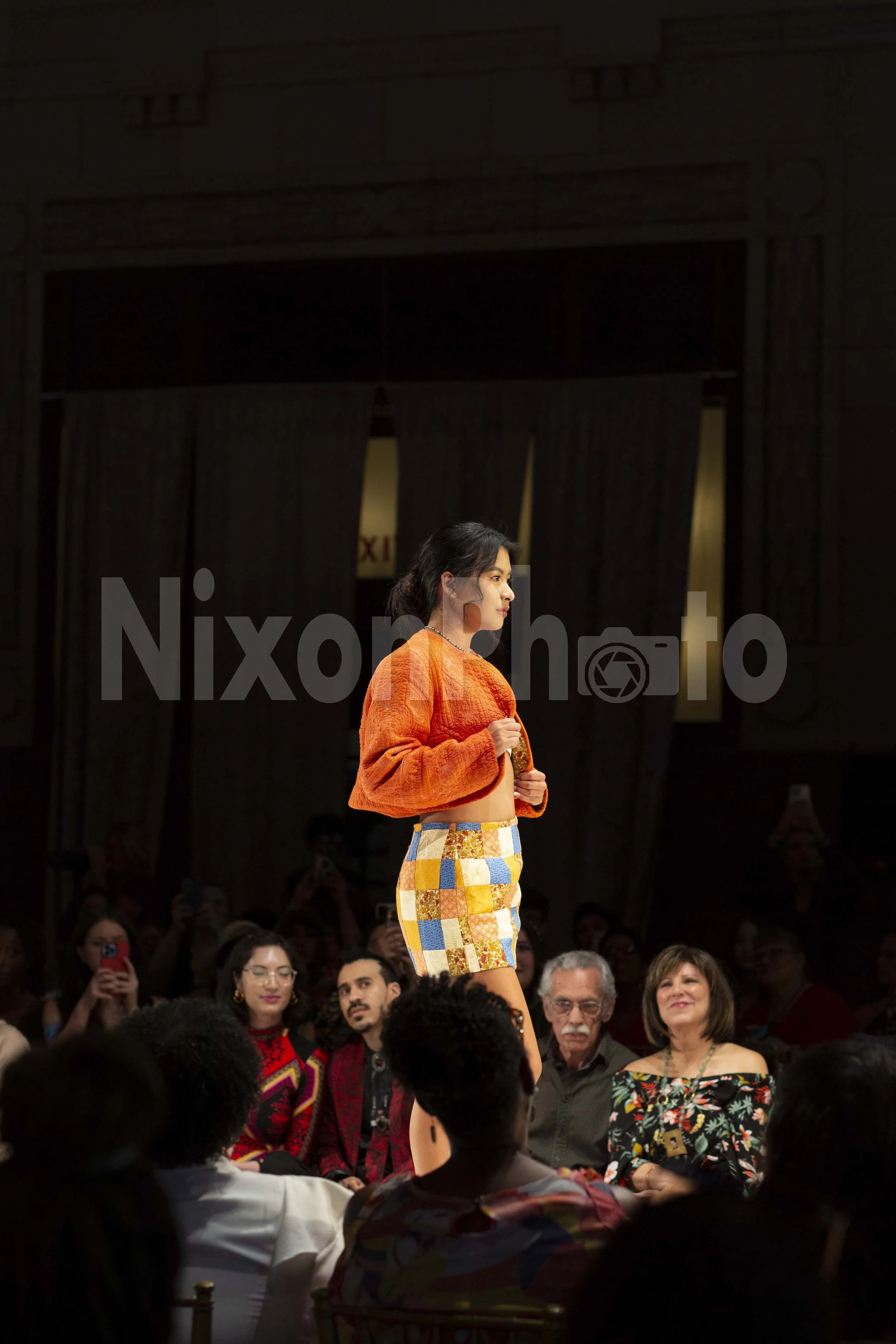 Model walking a runway wearing an orange jacket and patchwork skirt at a fashion show; audience seated and watching.