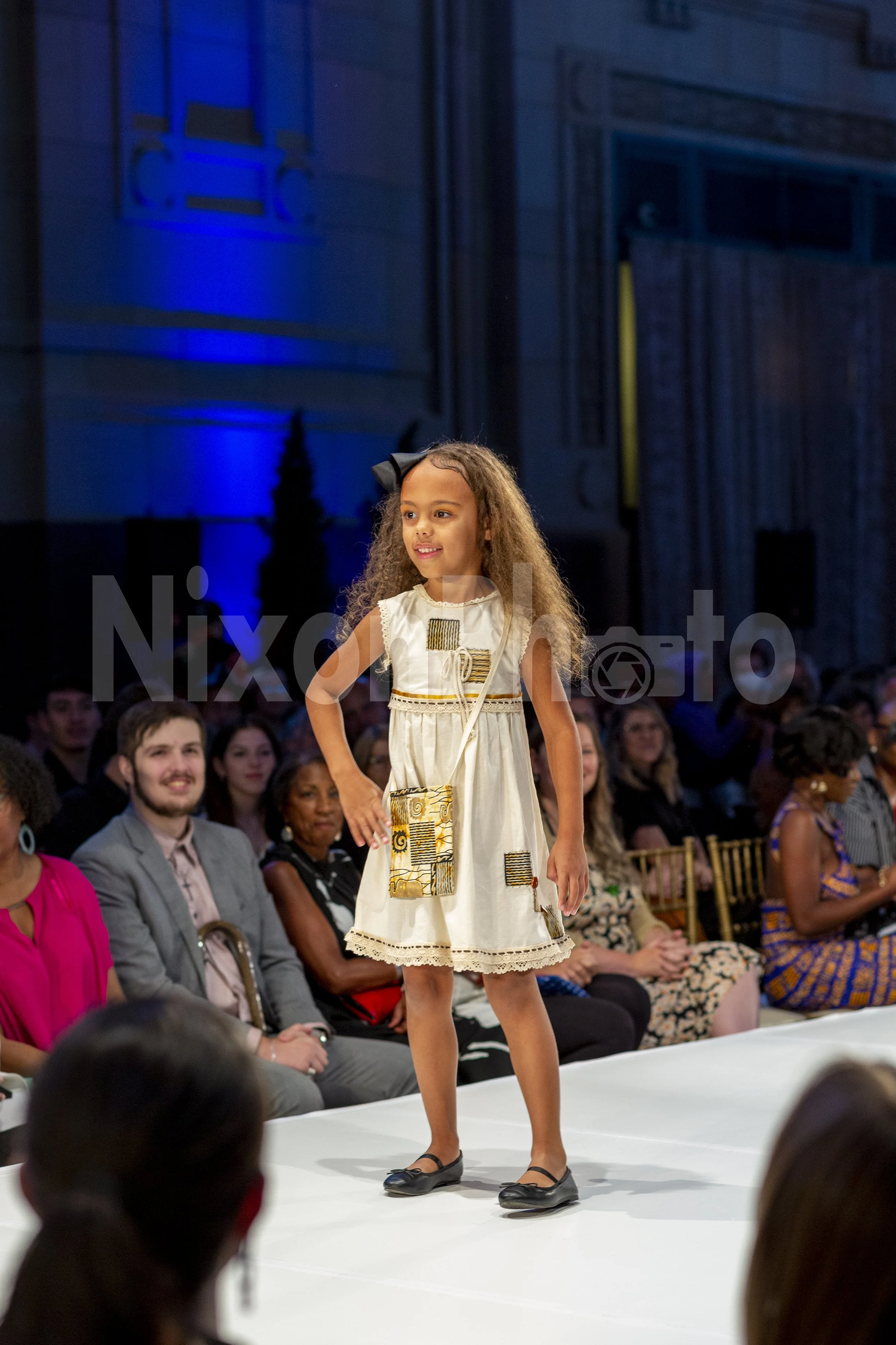 Young girl in a white dress walking on a runway during a fashion show, with seated audience watching.