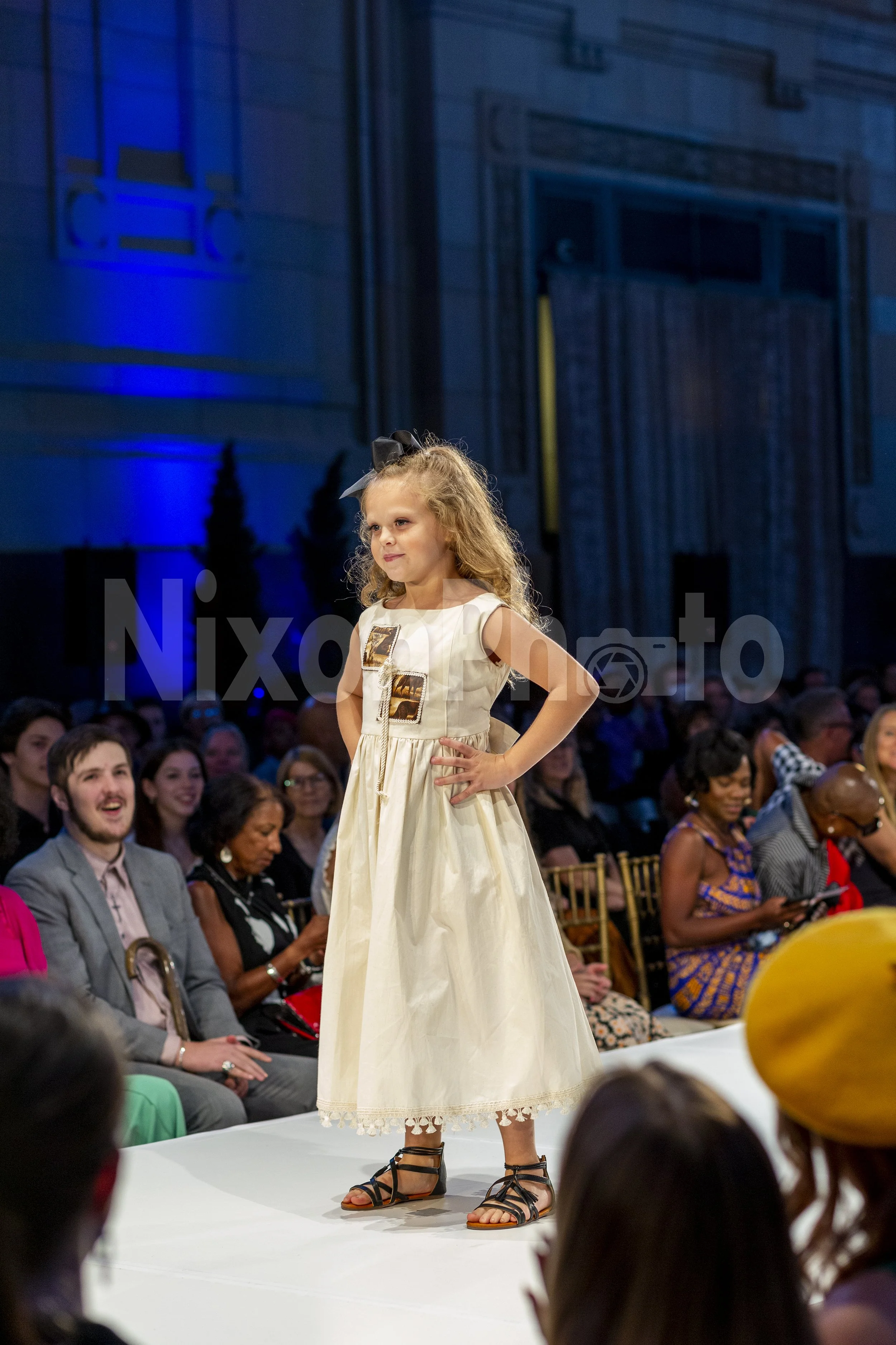 A young girl in a white dress walks on a fashion runway with an audience watching.