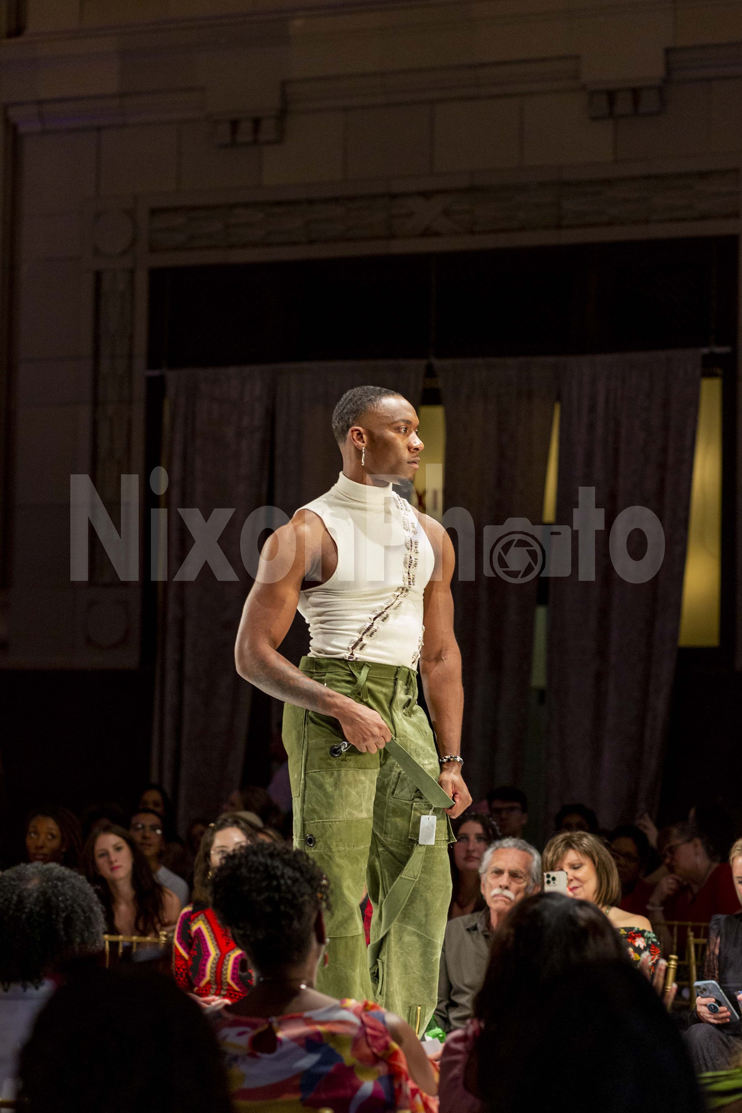Model walking on a runway wearing a sleeveless top and green pants, with audience seated in the background.