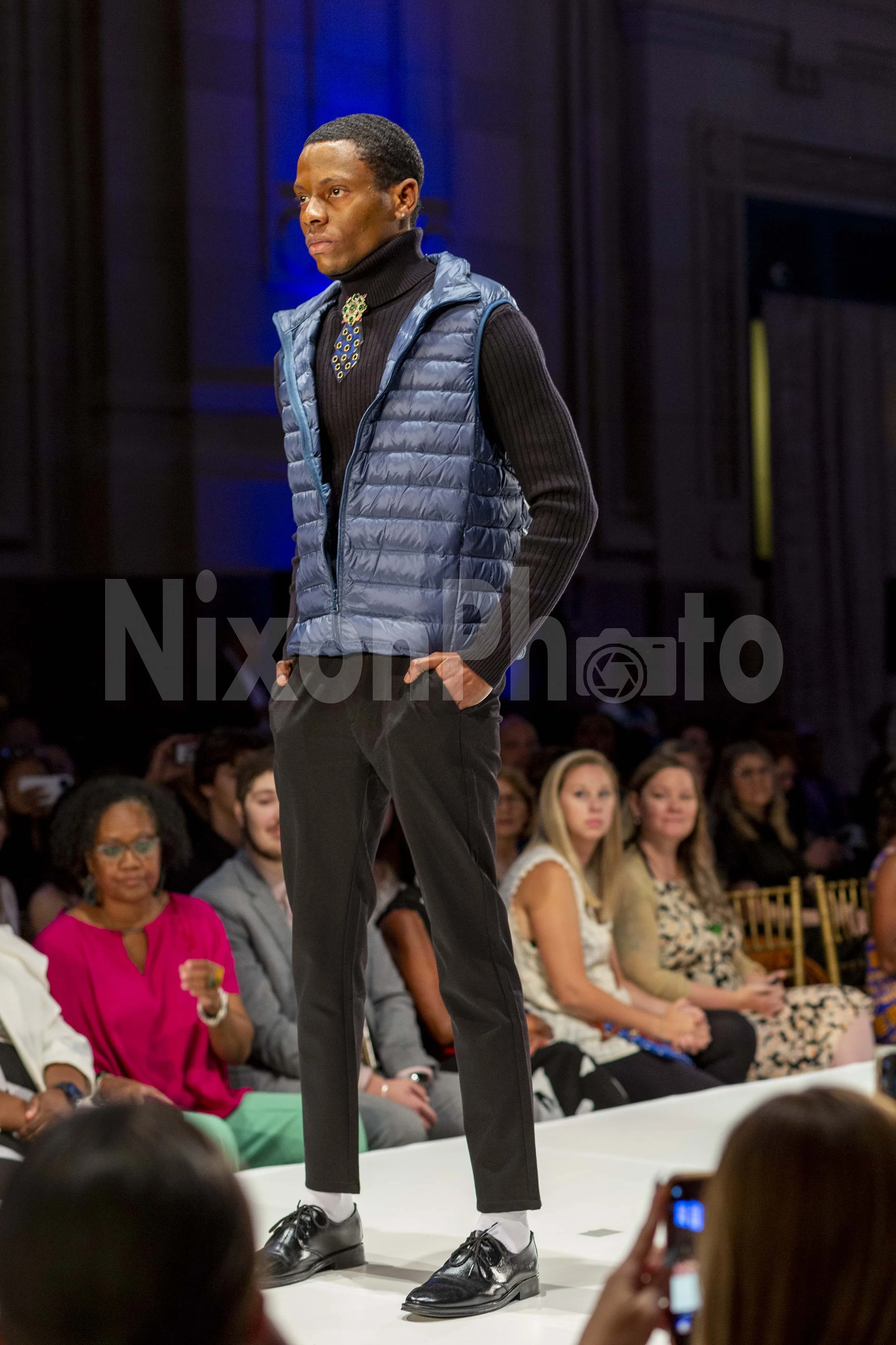 Model walking on a runway in blue puffer vest and black pants, with audience watching.