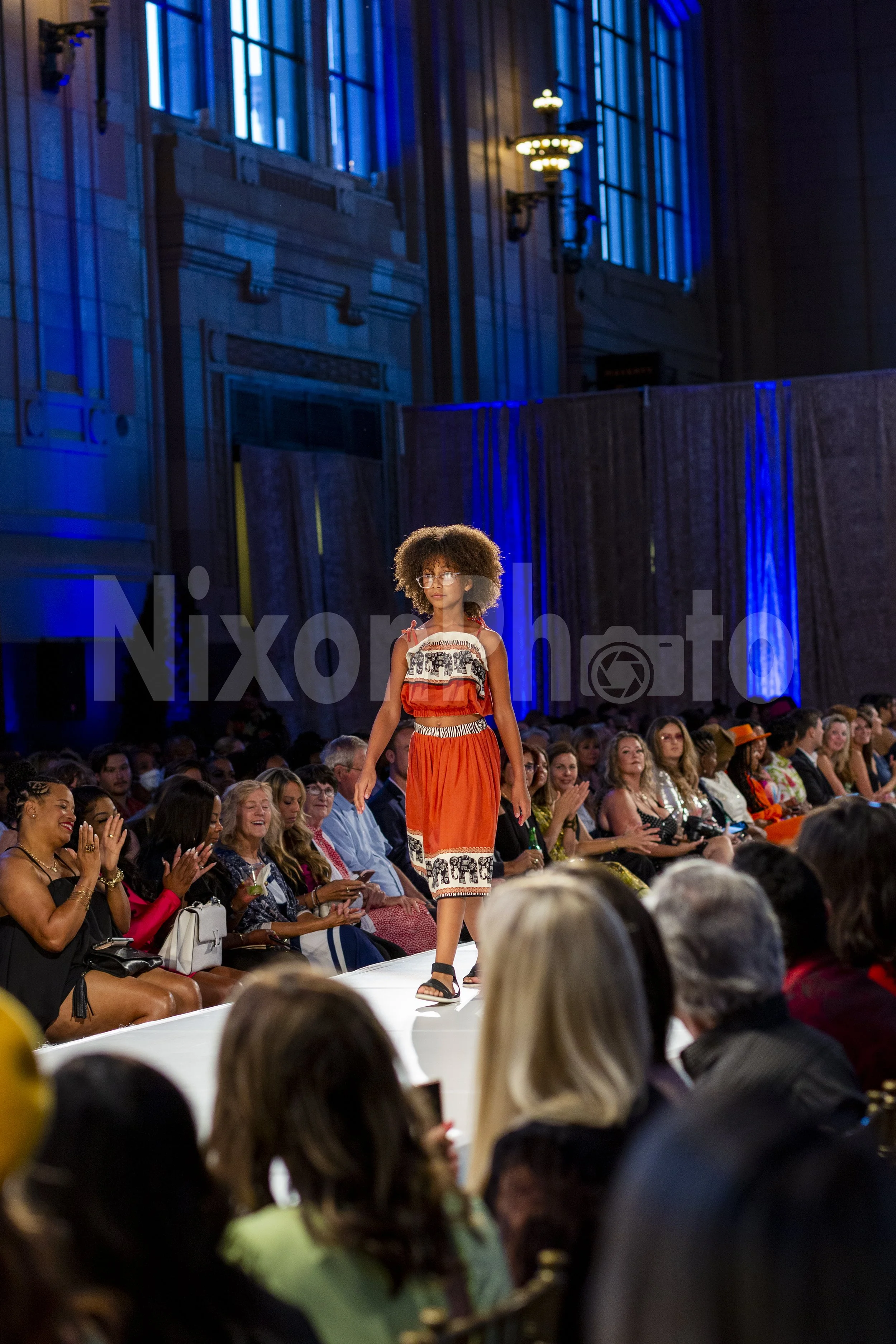 Fashion show with a model in an orange dress on a runway, audience applauding, and elegant venue with blue lighting.