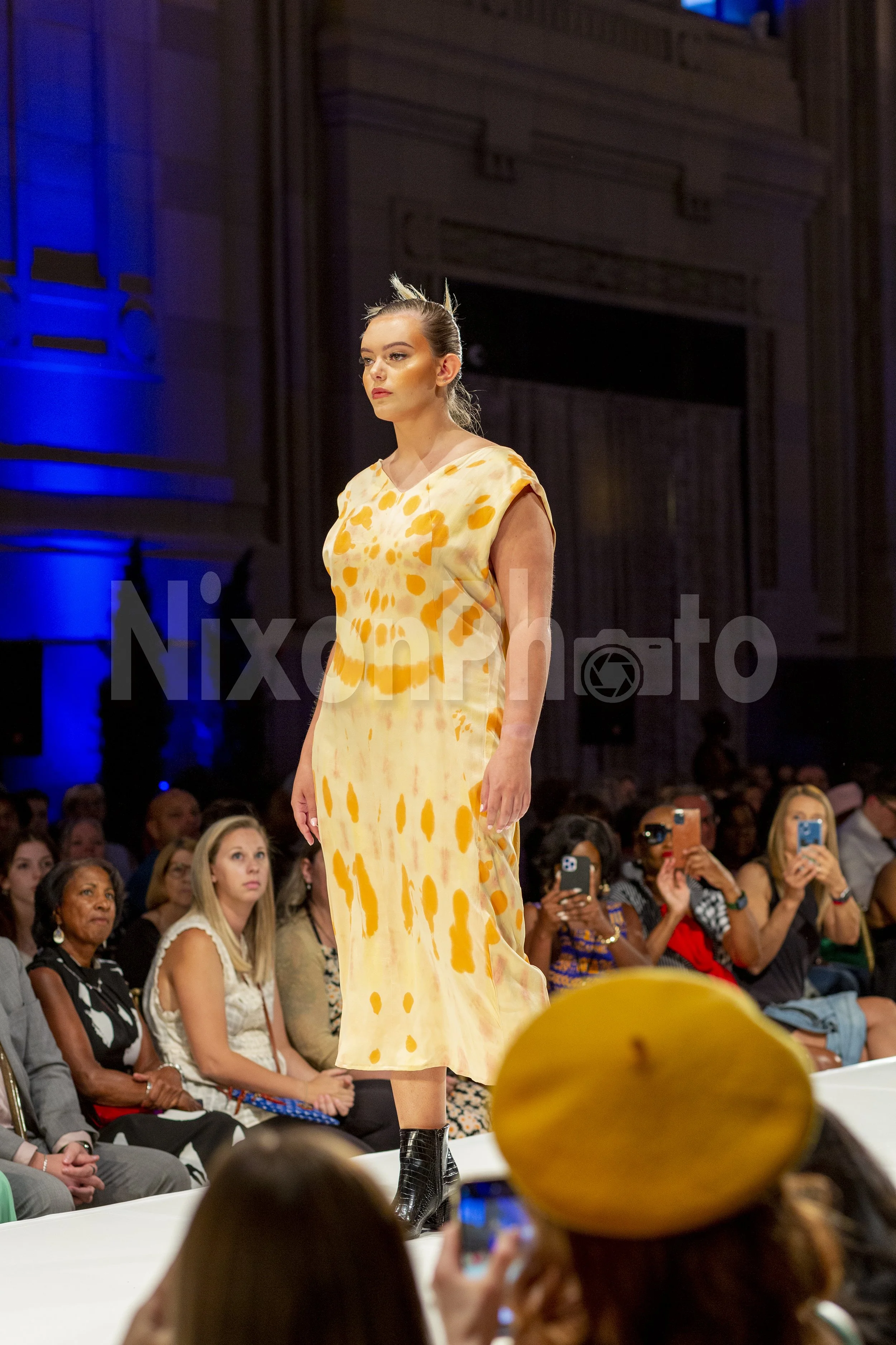 Fashion show with model in yellow patterned dress on runway, audience in background.