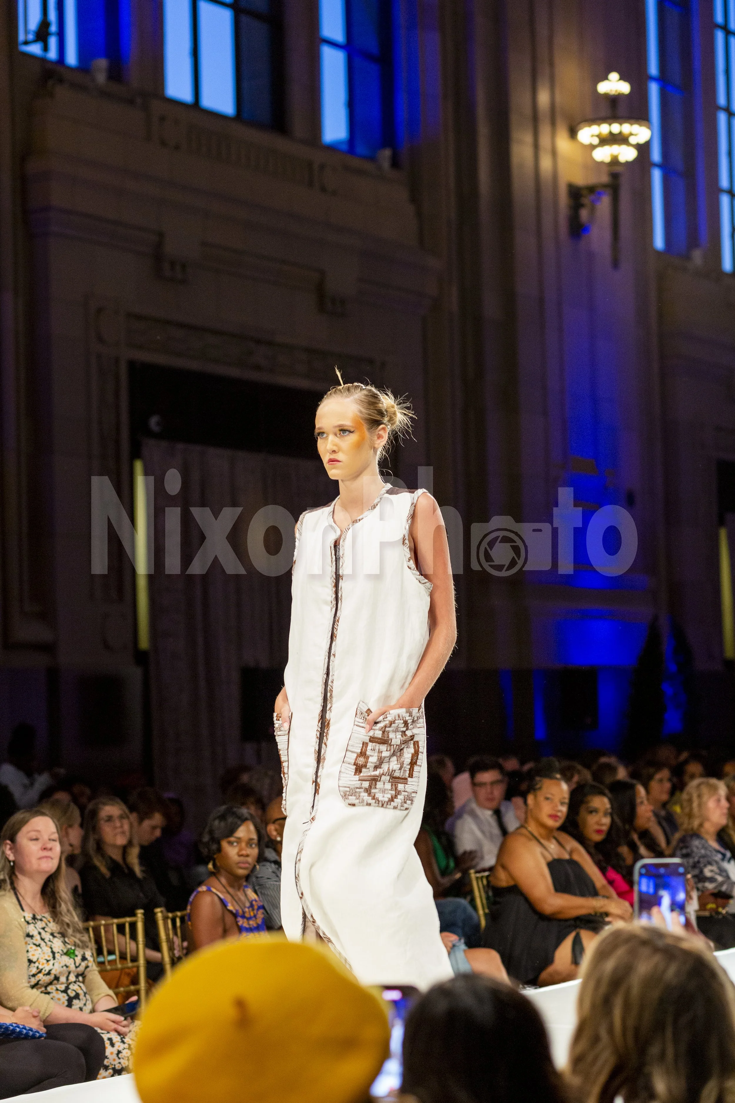 Model walking down a runway in a fashion show wearing a white dress with brown accents, audience seated along the sides in a dimly lit venue with blue lighting.