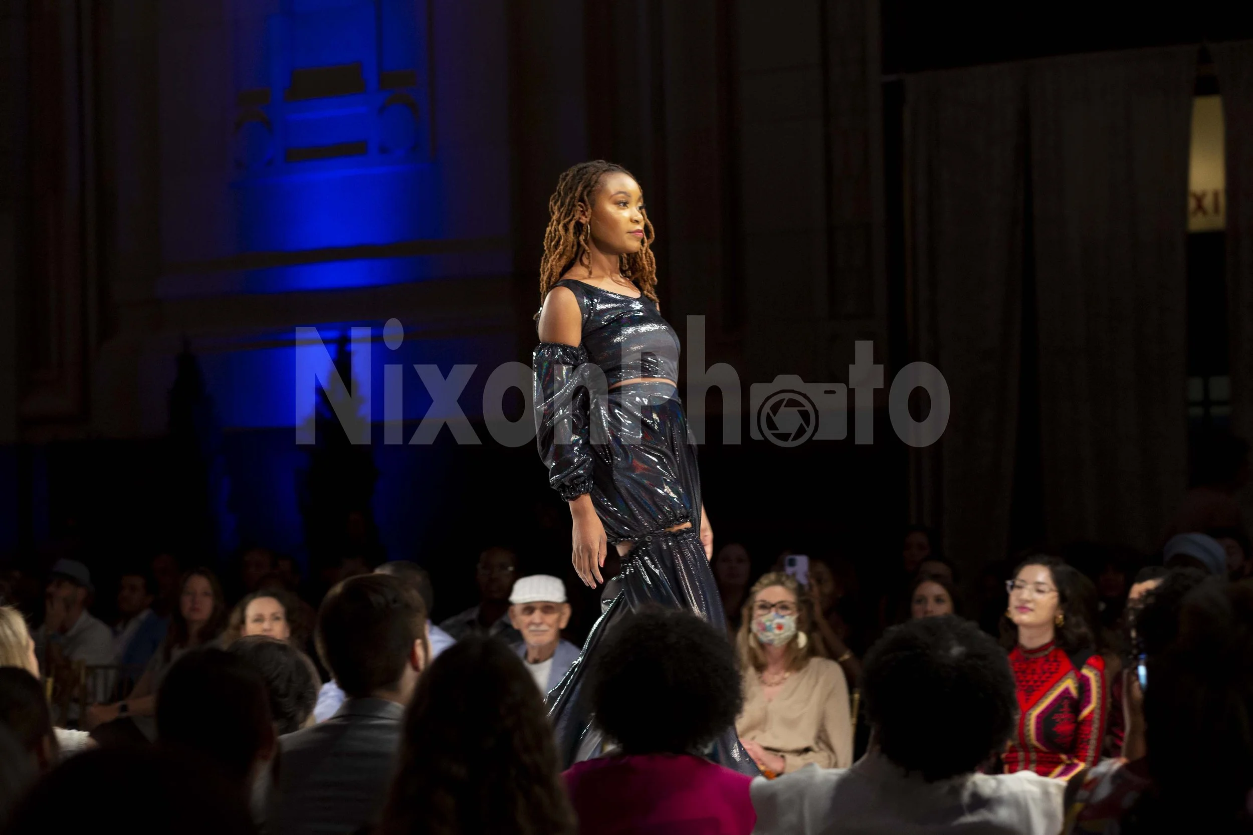 Model in black metallic dress walking on runway with audience seated on both sides in dimly lit room.