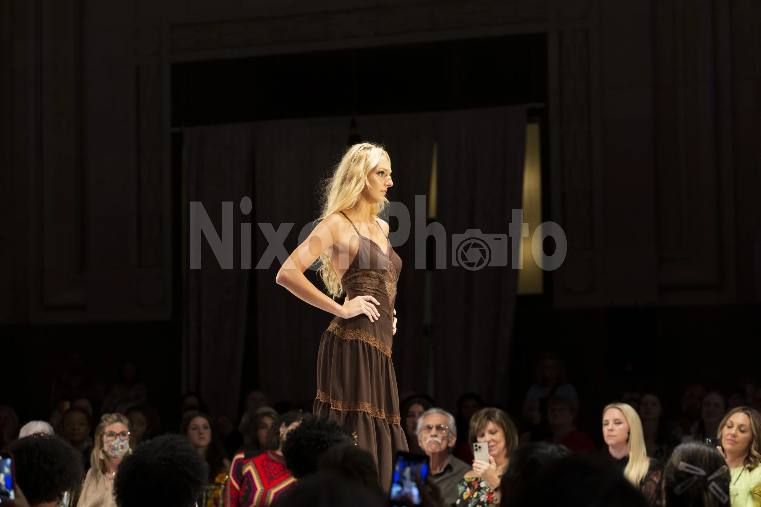 Model walking on a fashion runway, wearing a brown dress, with an audience watching.