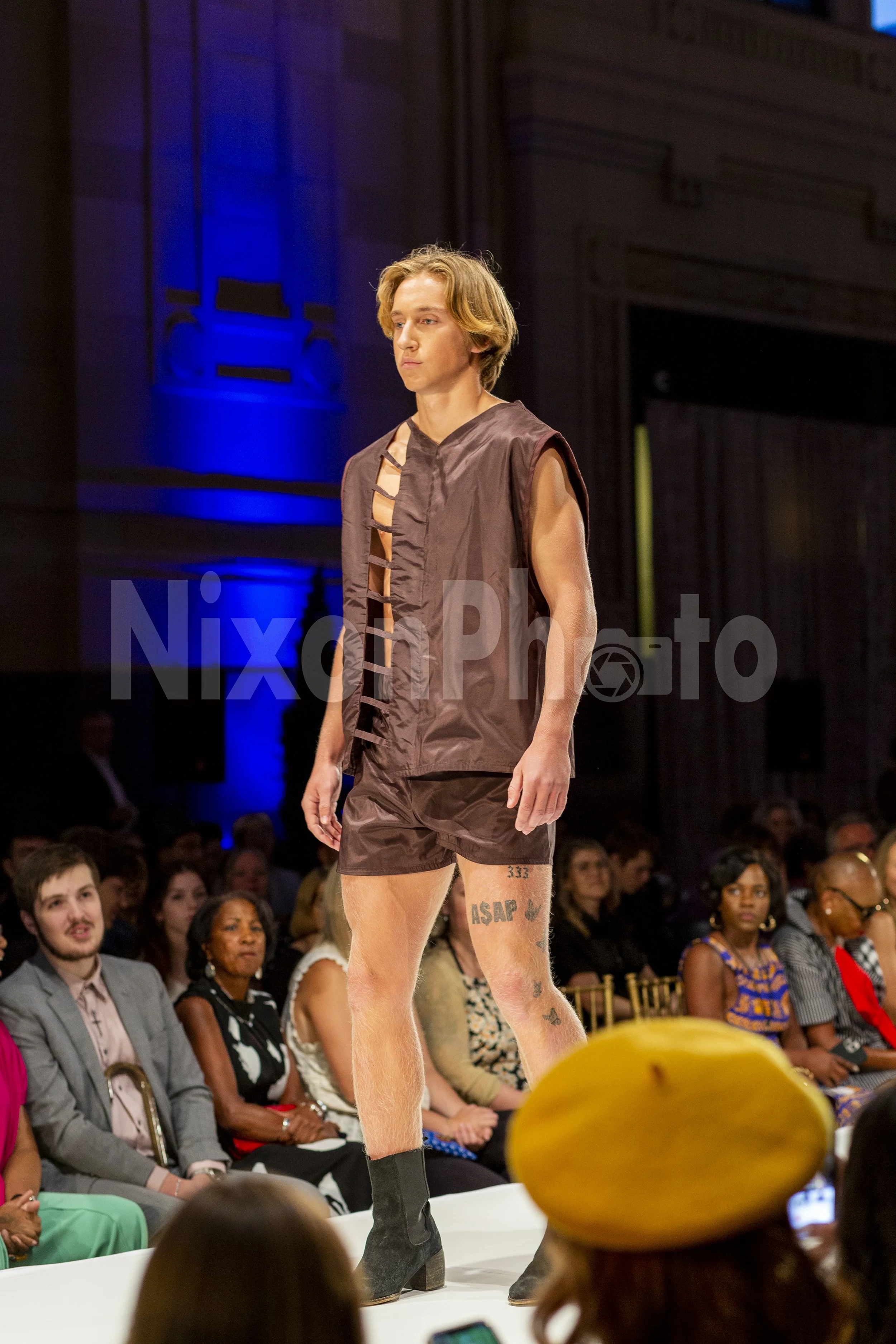 Male model wearing a sleeveless brown outfit walks on a fashion runway with seated audience in the background.