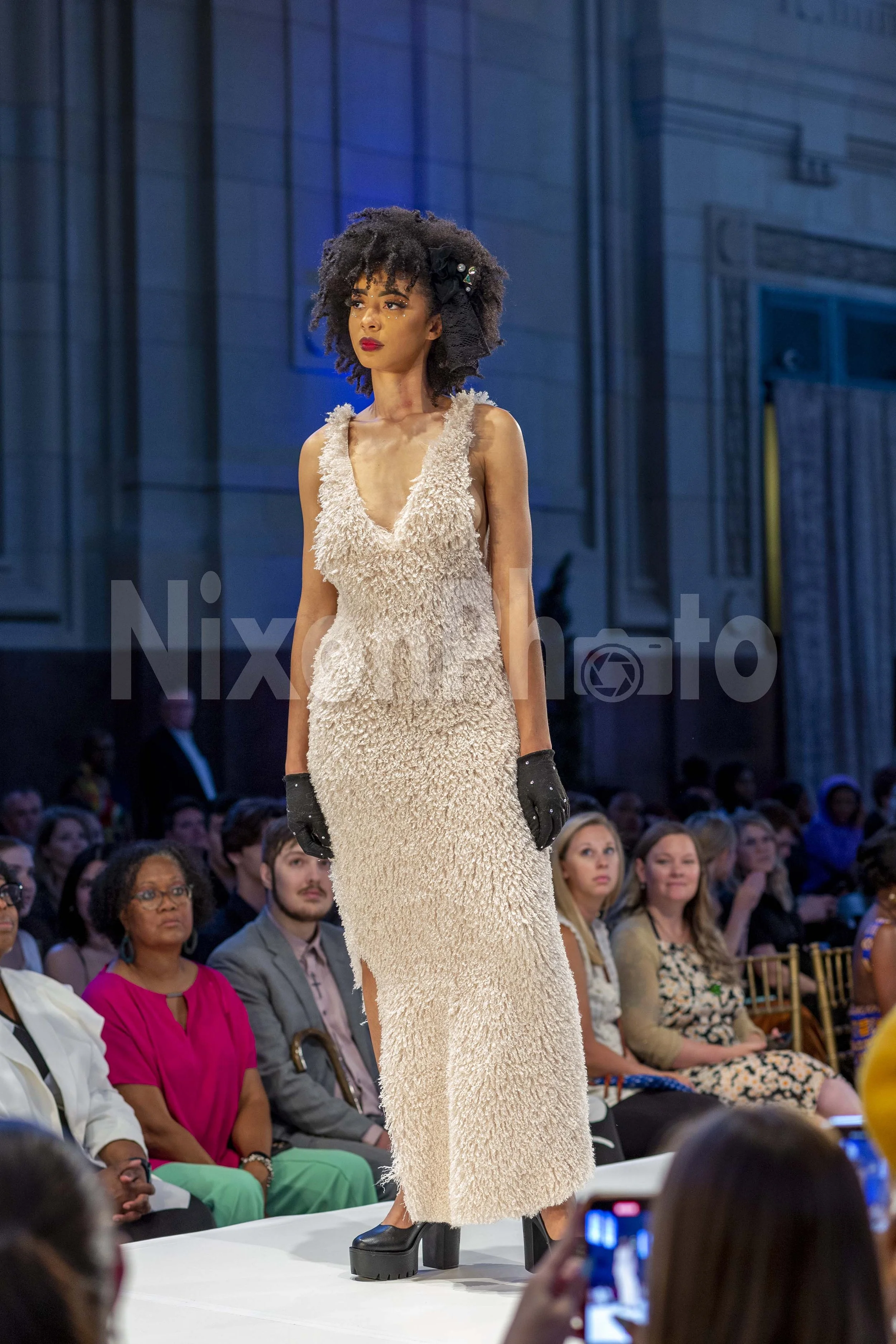 Fashion model walking on runway in a furry beige dress and platform shoes, surrounded by seated audience.