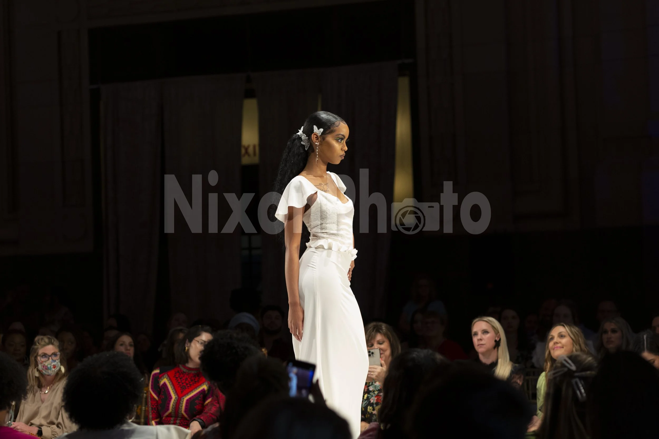 Model in white dress walking down a fashion runway with an audience watching.