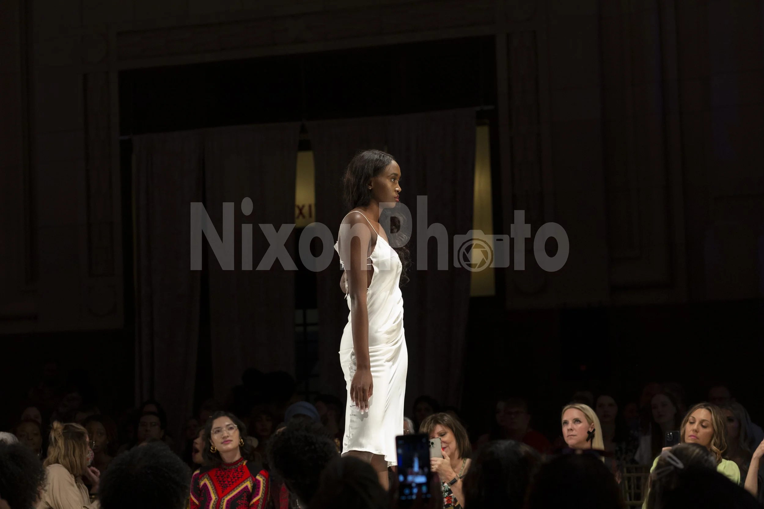 Model walking on a runway at a fashion show, wearing a white dress, with an audience seated in the background.