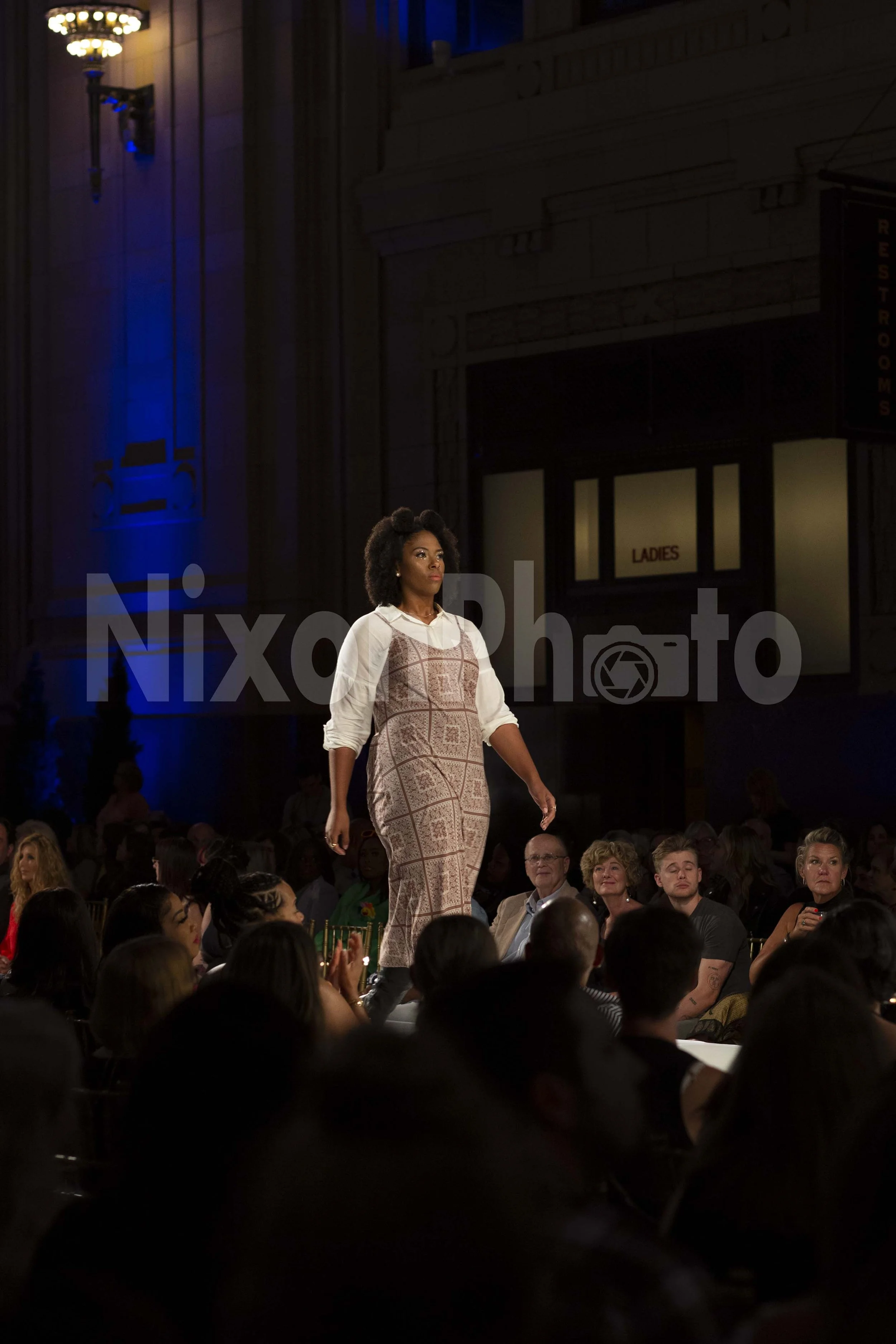 Fashion show runway with a model wearing a patterned dress, audience watching in the background, blue lighting.