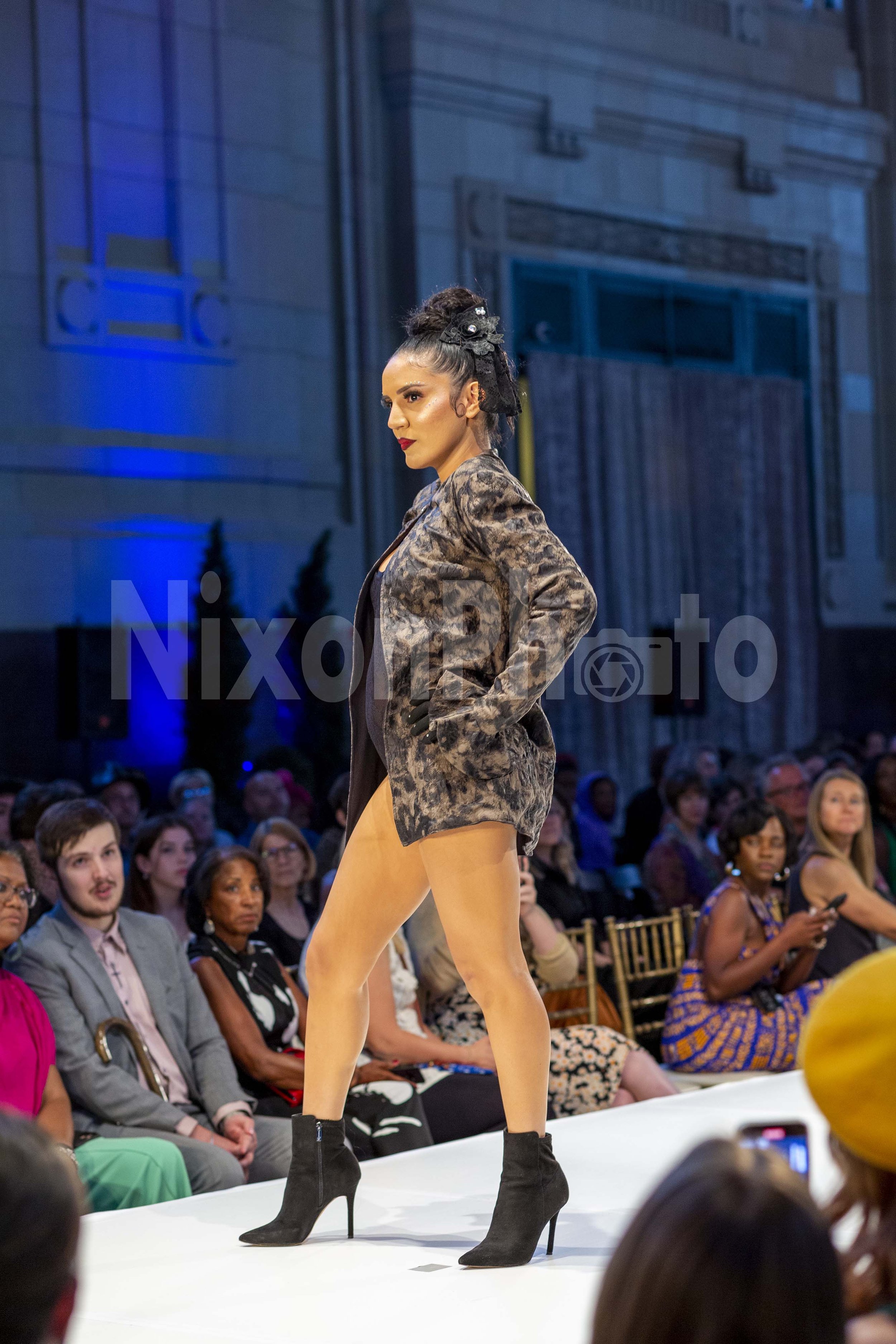 Model walking on runway in fashion show with audience seated and watching.