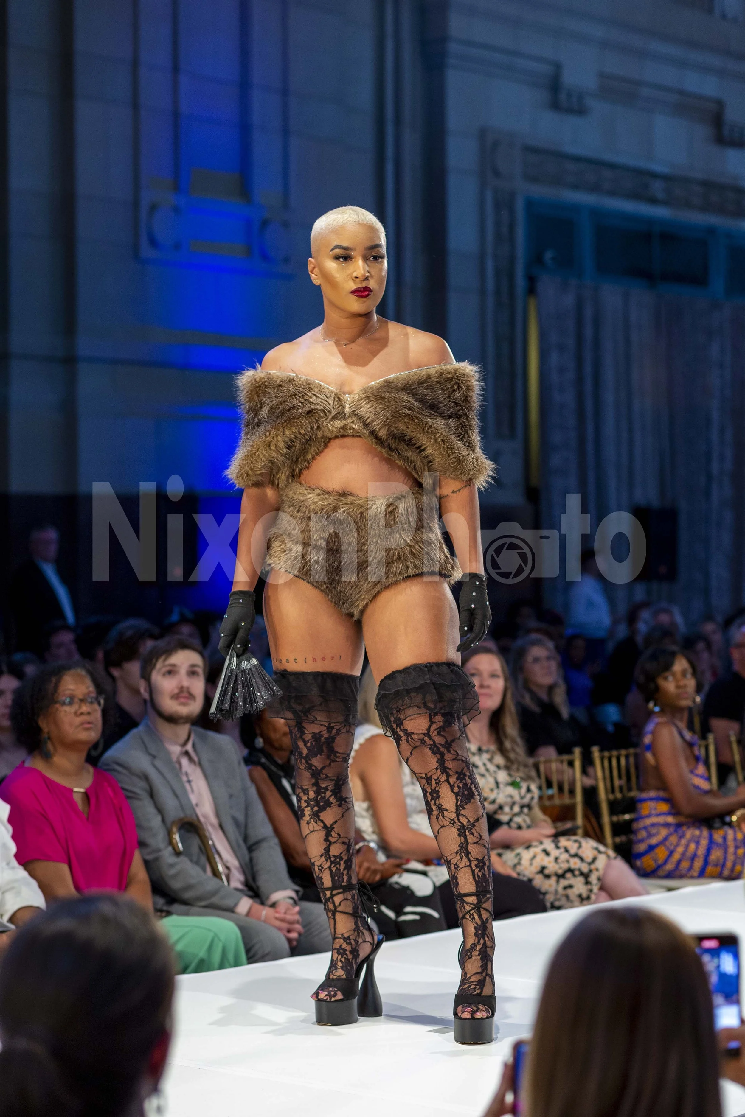 Model wearing fur outfit with lace boots on runway at fashion show, audience in background.