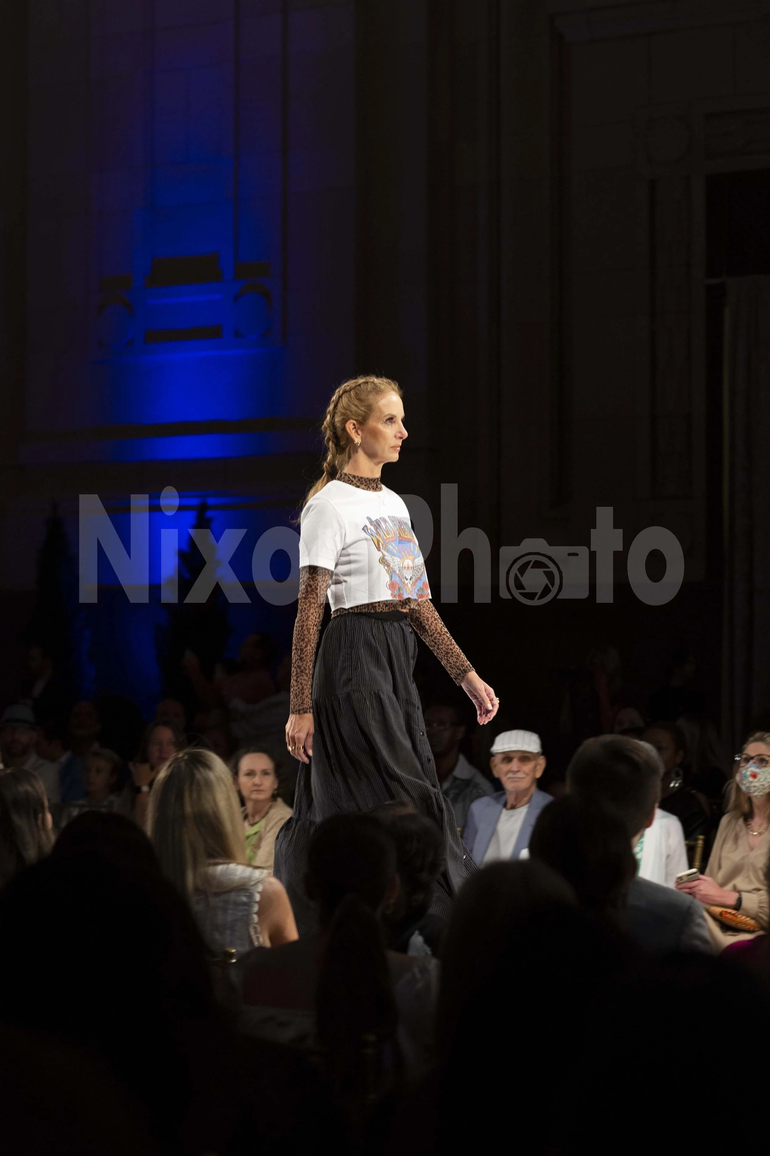 Model walking down a runway wearing a graphic t-shirt and long black skirt at a fashion show with spectators seated on either side.