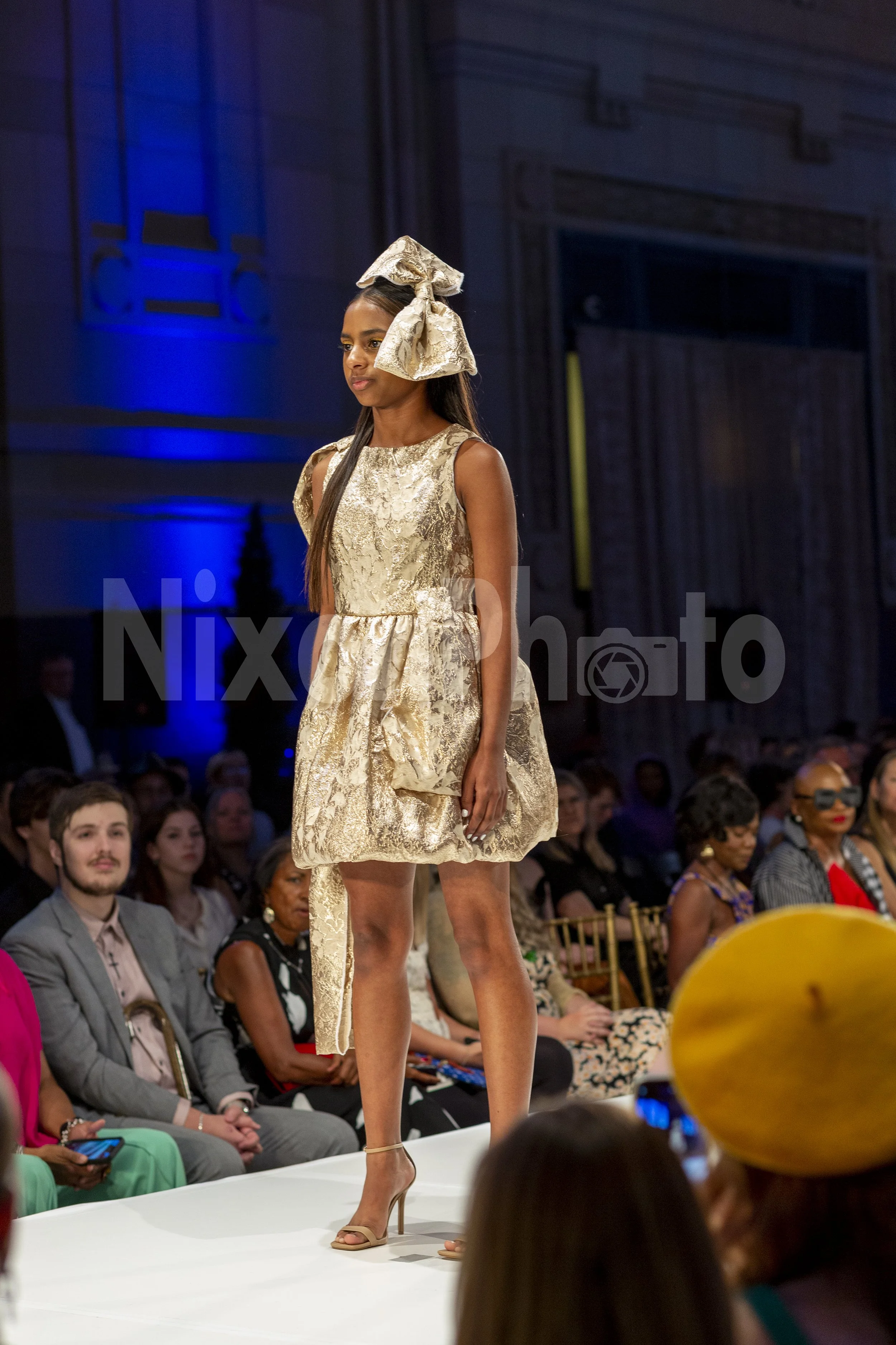 Model wearing a gold dress with a large bow on the runway at a fashion show.