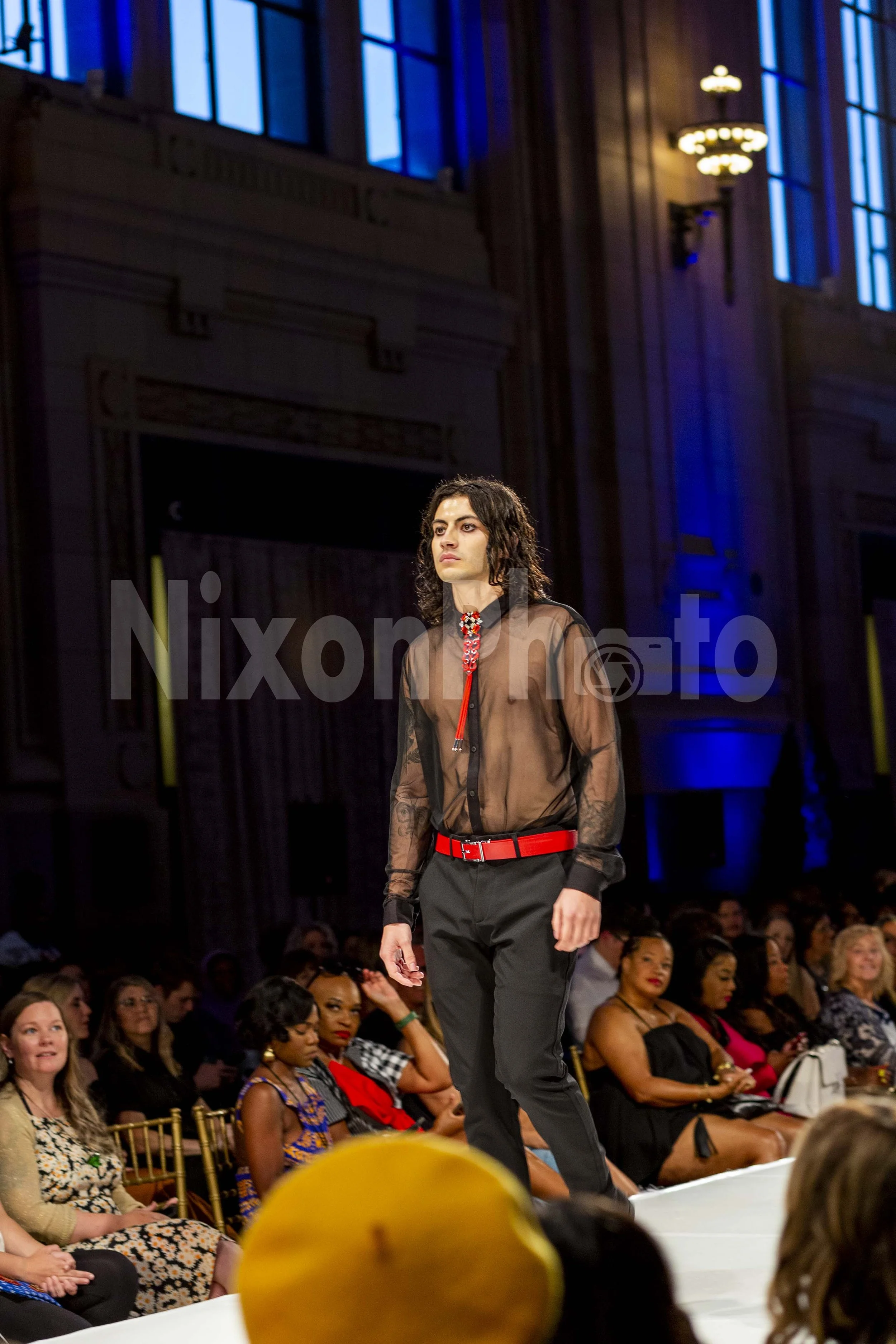 Model walking on a runway wearing a sheer black shirt and red accents with an audience watching in a large hall.