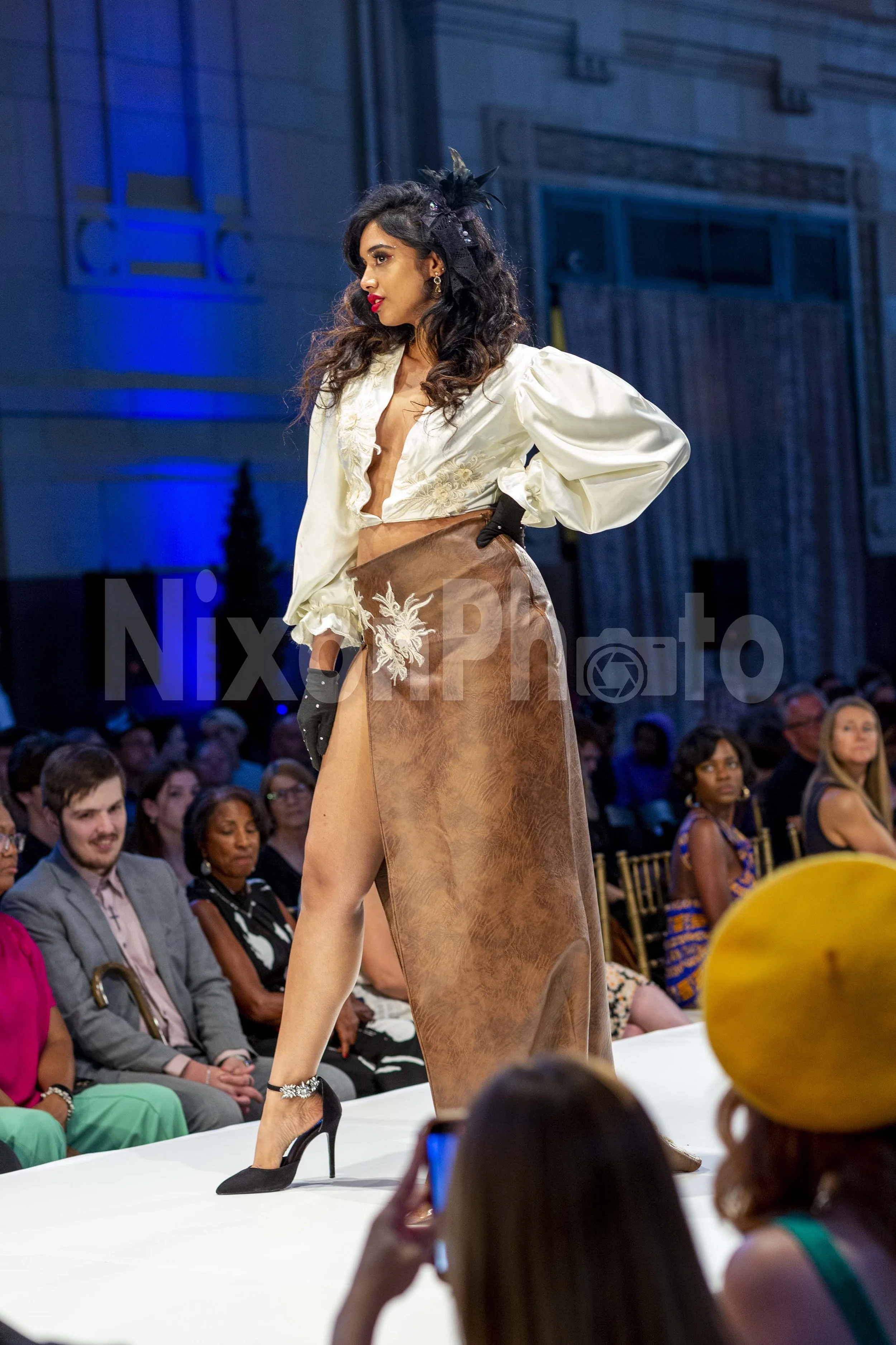 Fashion model walking on a runway wearing an elegant outfit with a long brown skirt, a white blouse, and black heels. The audience is seated on either side of the runway.